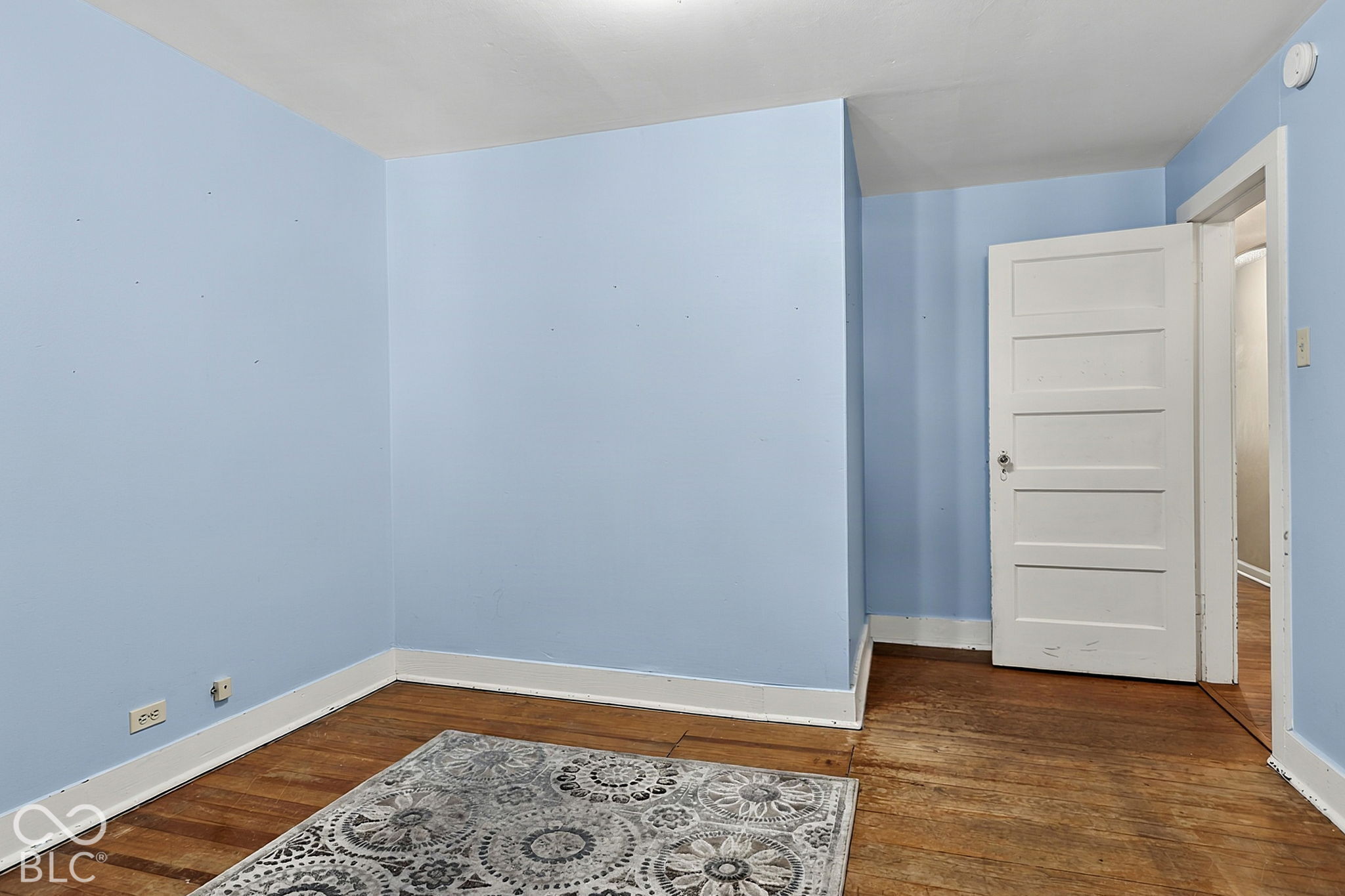 Empty room with pale blue walls, hardwood floors, and an open white door.