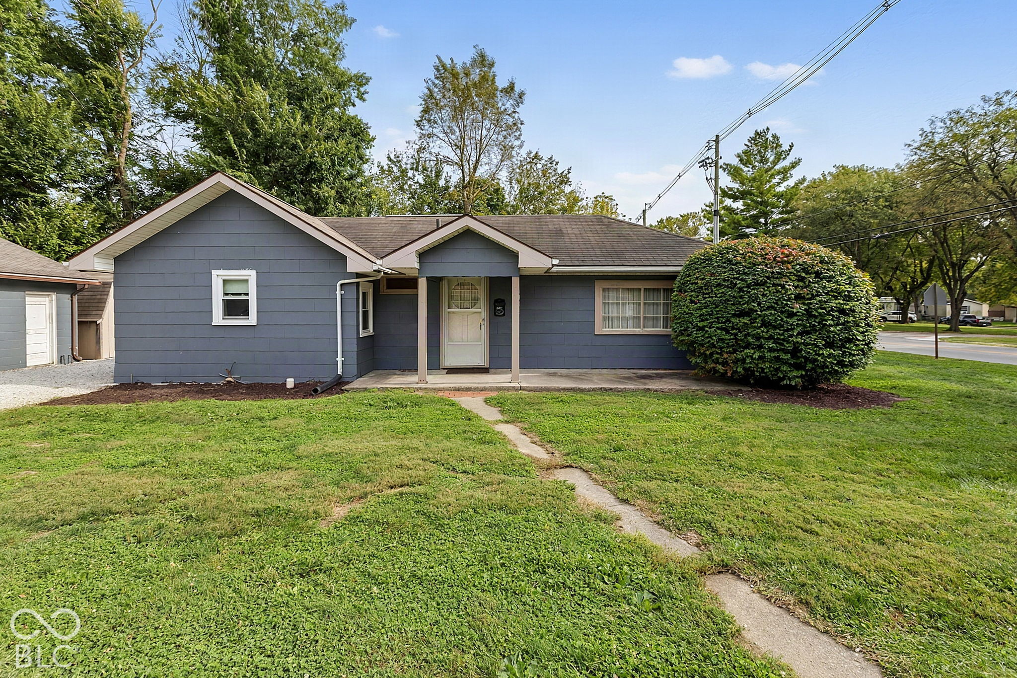 Gray single-story home with a covered entry, large rounded shrub, and concrete path across the front lawn.