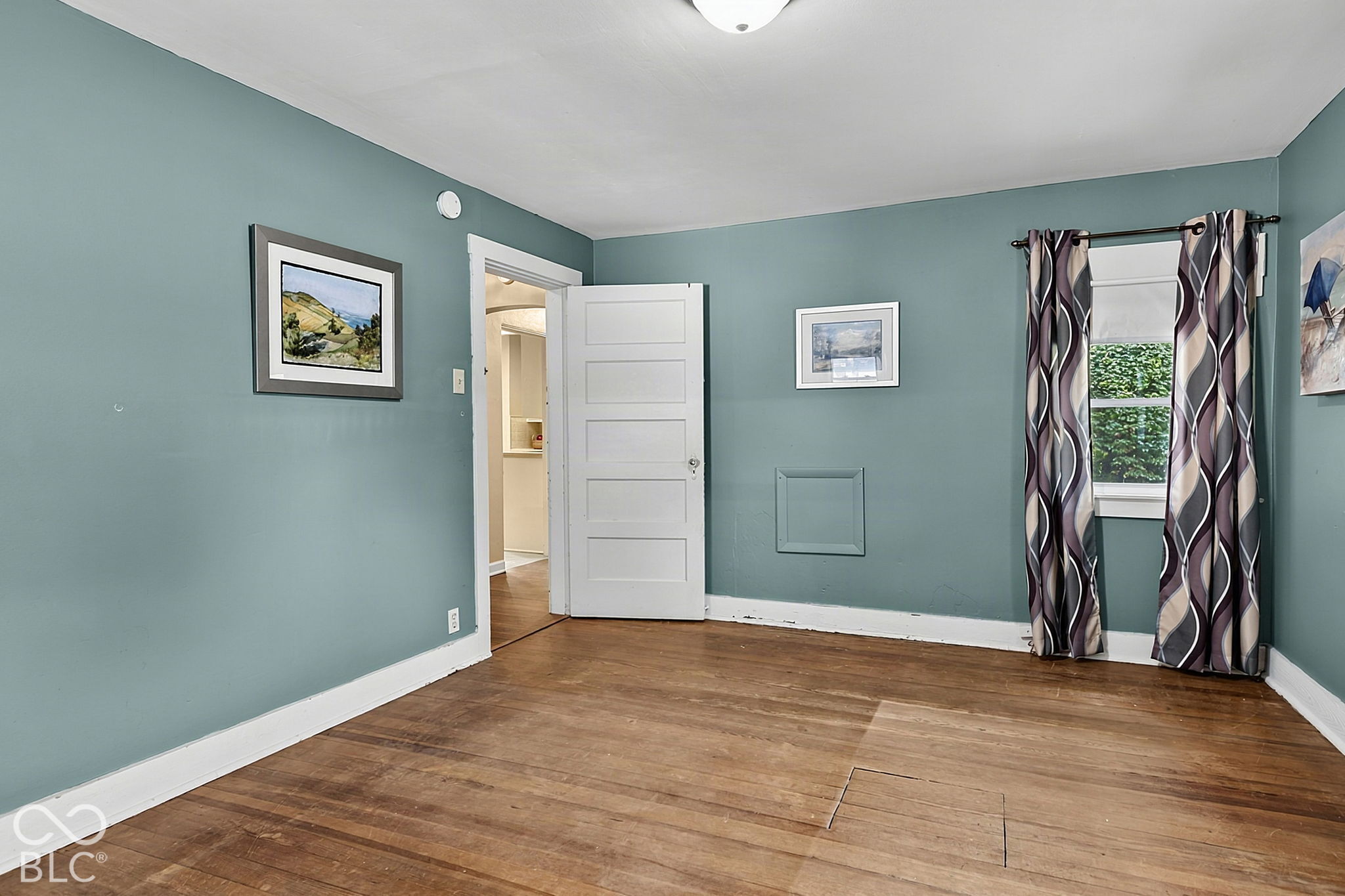 Unfurnished room with blue-green walls, hardwood floors, patterned curtains, and an open doorway.