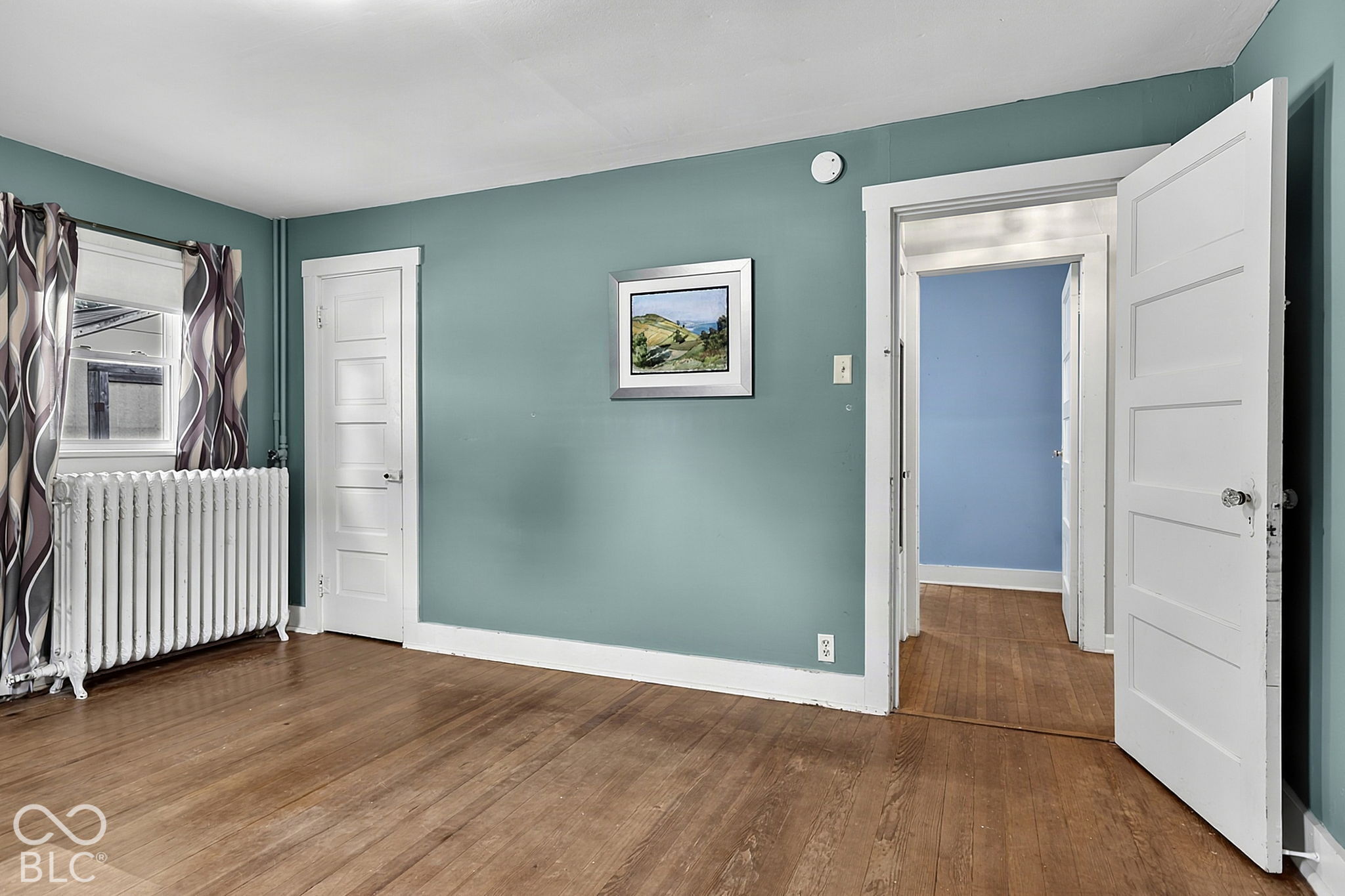 Empty teal room with hardwood floors, a white radiator, and an open doorway to a blue room.