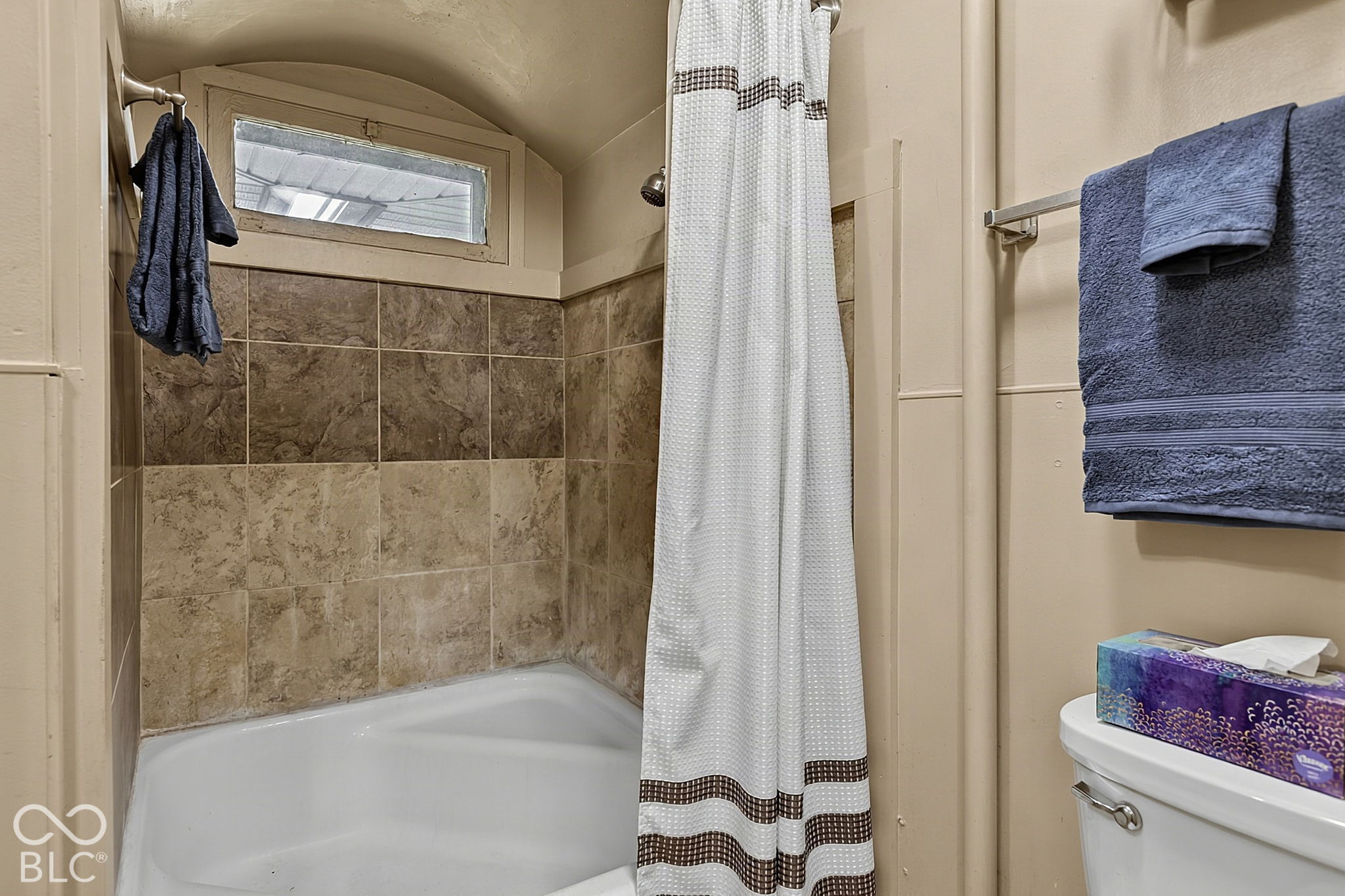 Bathroom with a tiled tub and shower, high window, white curtain, and blue towels.