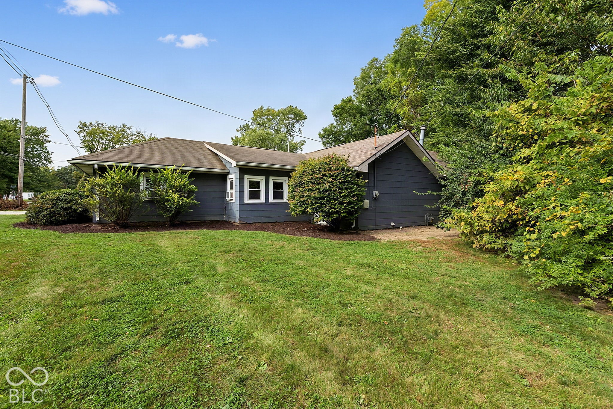 Single-story dark blue-gray house behind a broad lawn, with mature trees and shrubs.