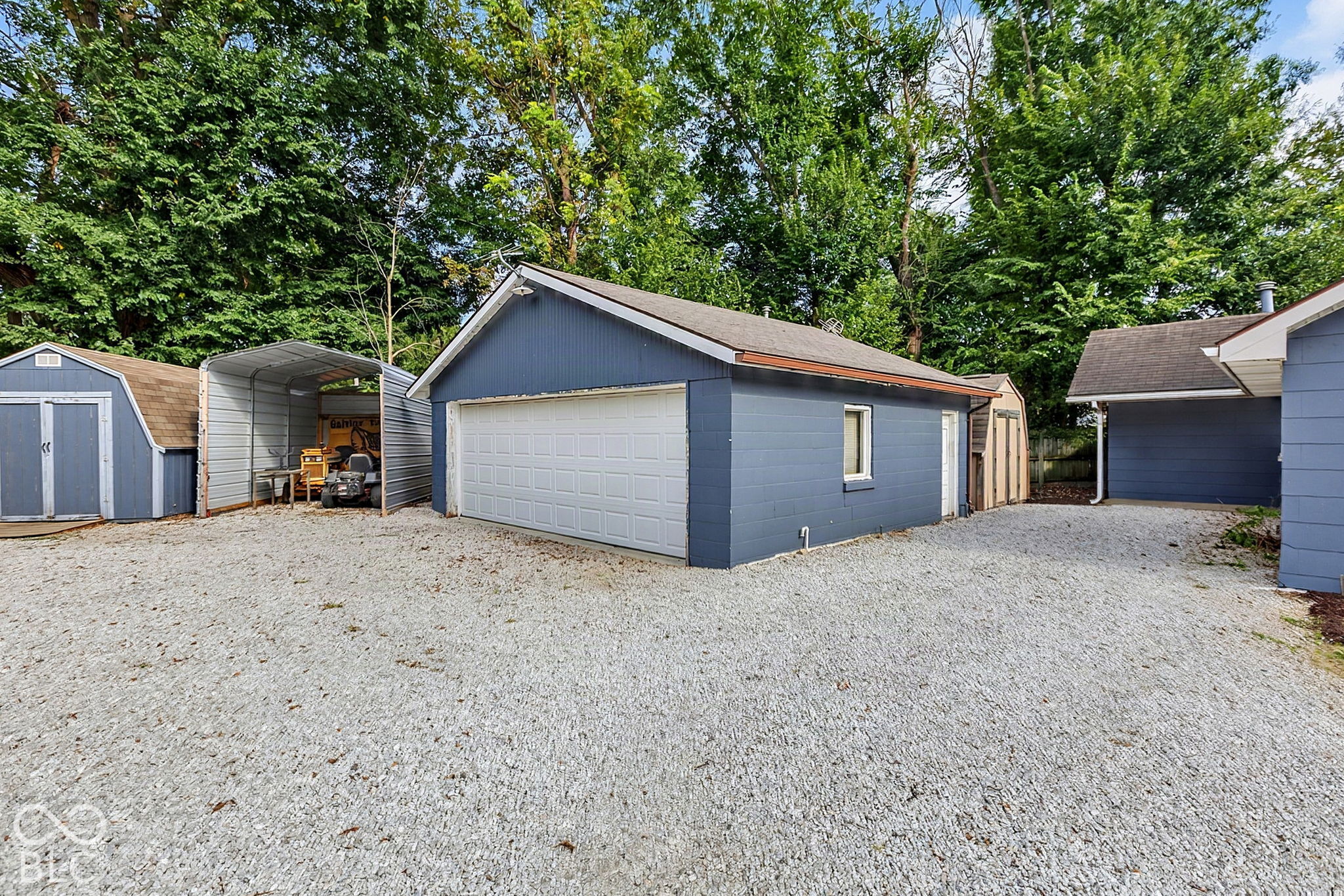 Blue detached garage with a white double door, gravel yard, and several nearby storage sheds.