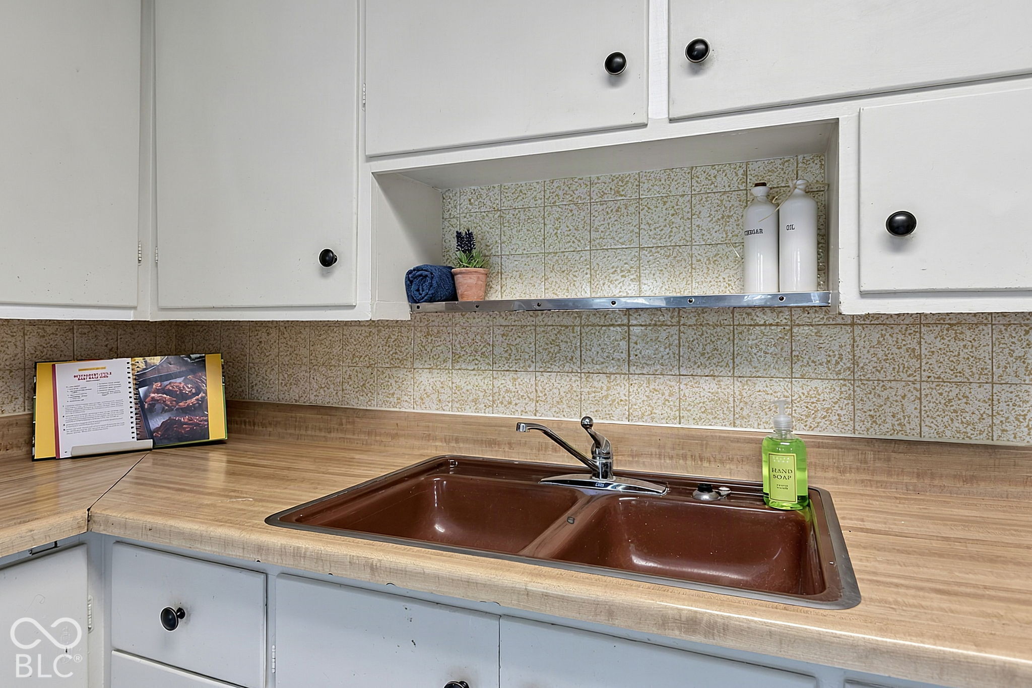 Kitchen sink with brown double basin, wood-look countertop, white cabinets, and patterned tile backsplash.