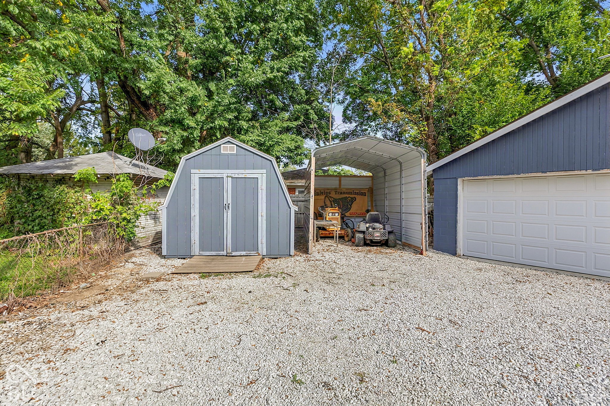 Gravel backyard with a gray storage shed, metal carport, and blue detached garage beneath mature trees.