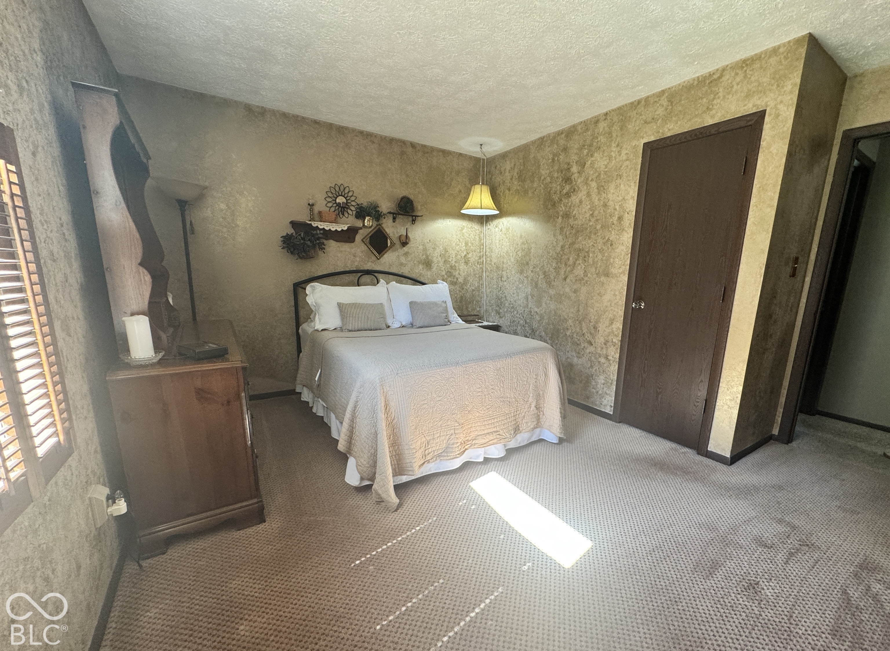 Furnished bedroom with a beige-quilted bed, textured tan walls, and sunlight across the carpet.