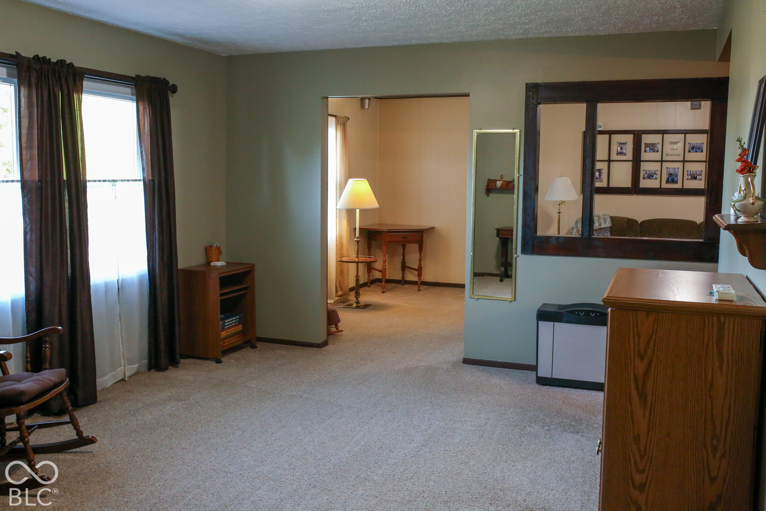 Carpeted room with a large curtained window, wooden cabinet, and an opening to a sitting area.