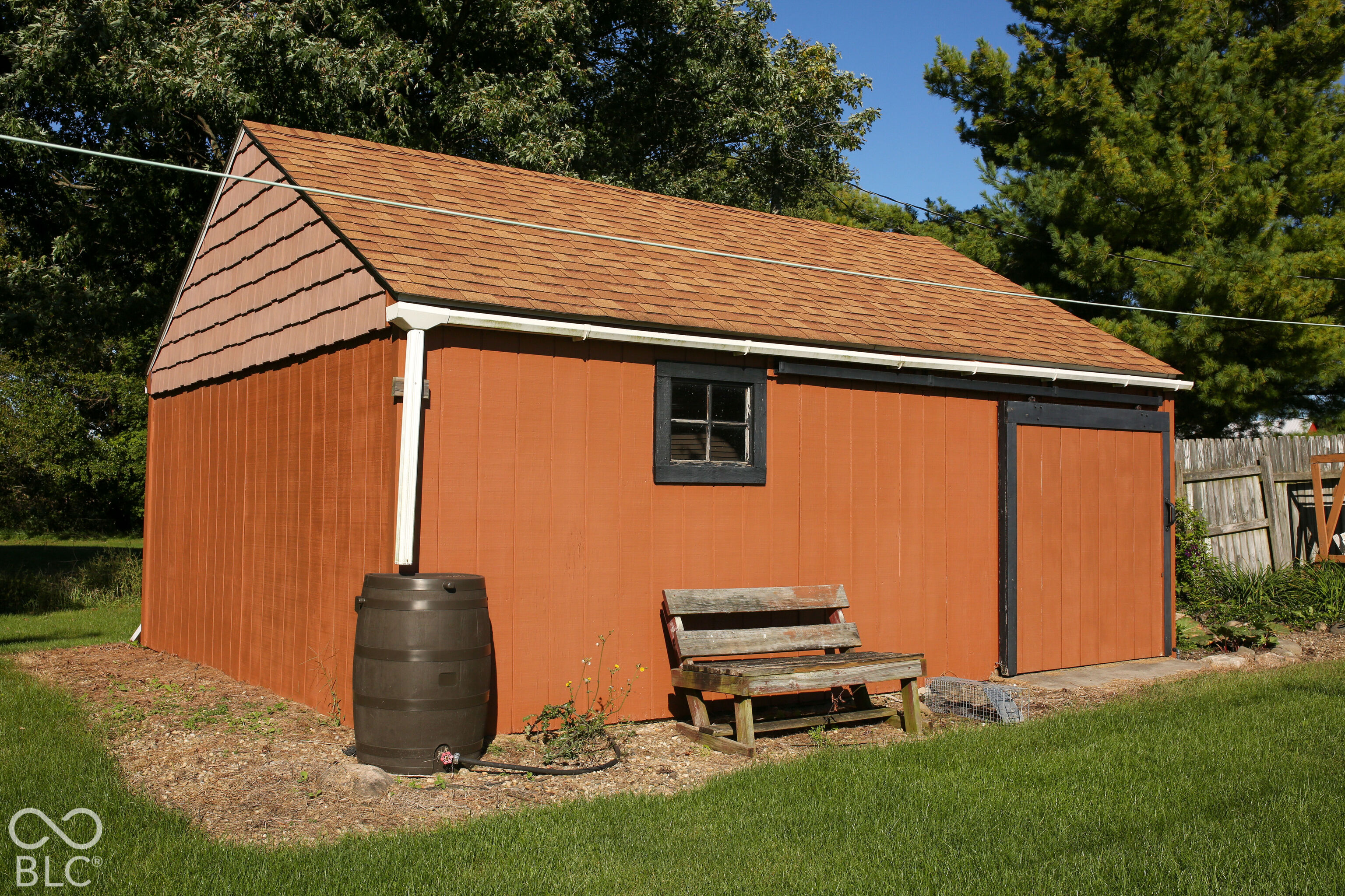 Red-orange backyard shed with a rain barrel and weathered wooden bench.