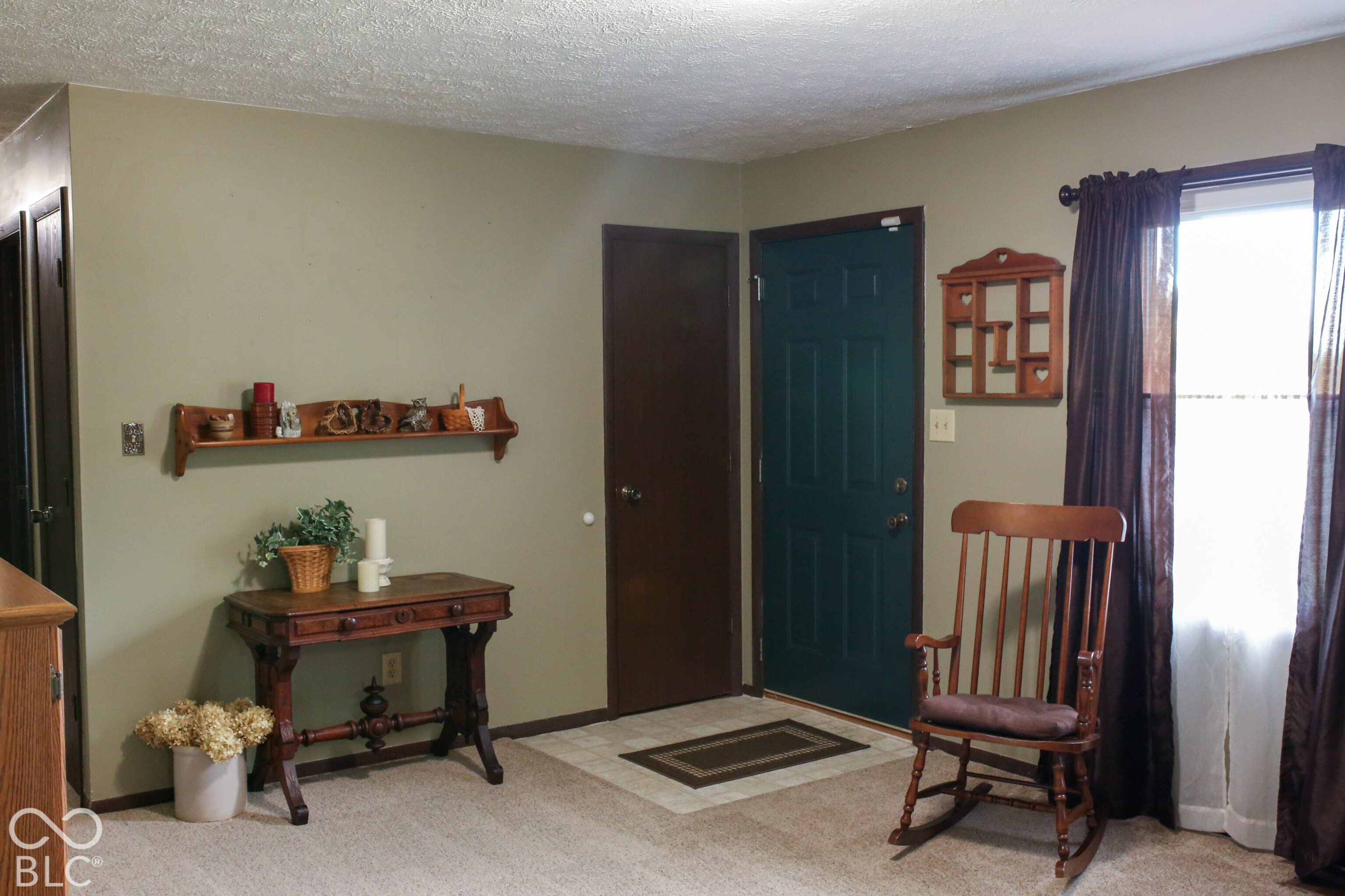 Furnished entryway with a dark teal front door, wooden console table, and rocking chair by a curtained window.
