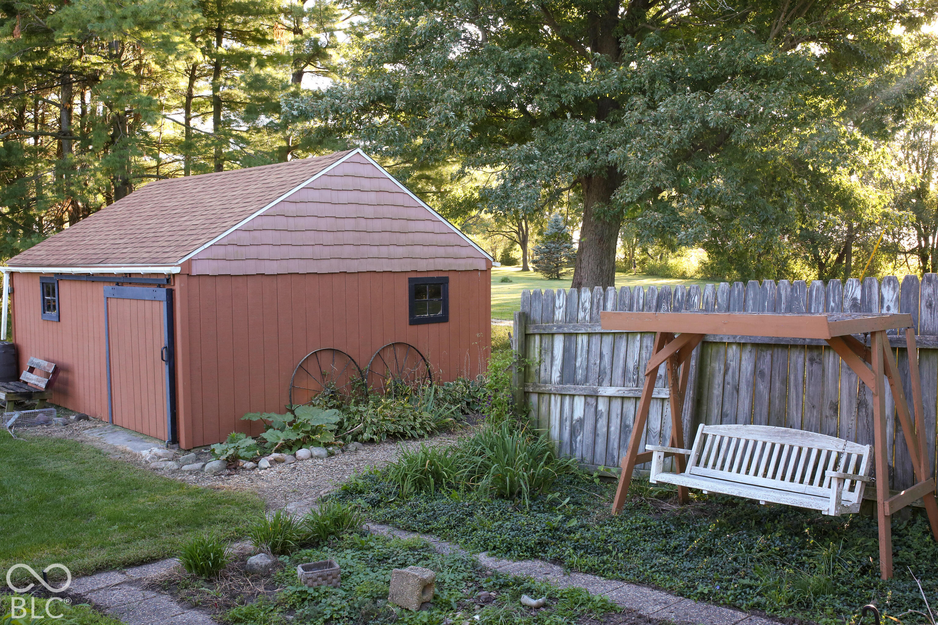 Red-brown backyard shed beside a wooden fence and garden, with a porch swing in front.