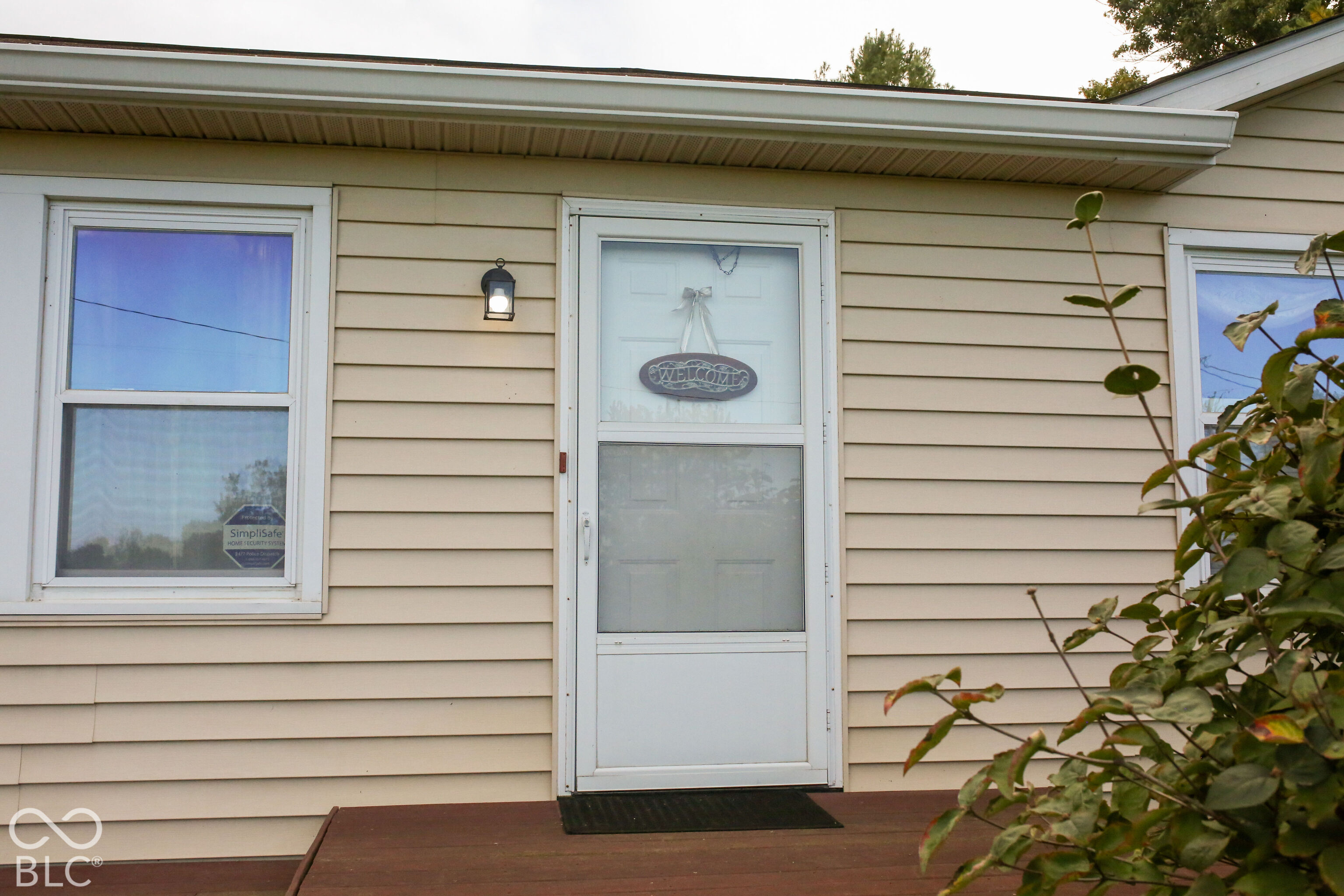 Beige-sided home entrance with a white storm door, welcome sign, and wooden porch.