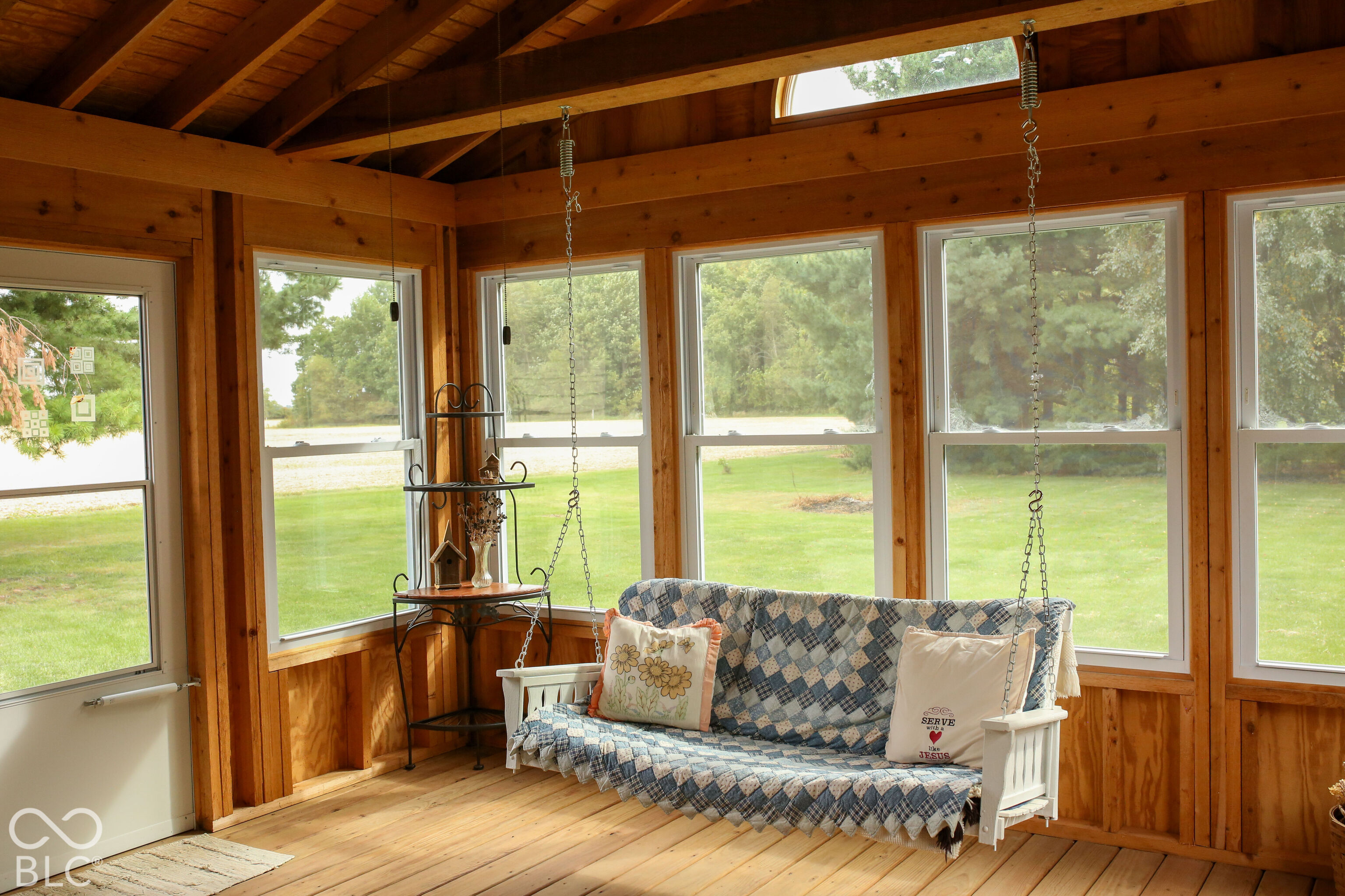 Wood-paneled sunroom with a quilt-covered hanging swing and large windows overlooking the lawn.