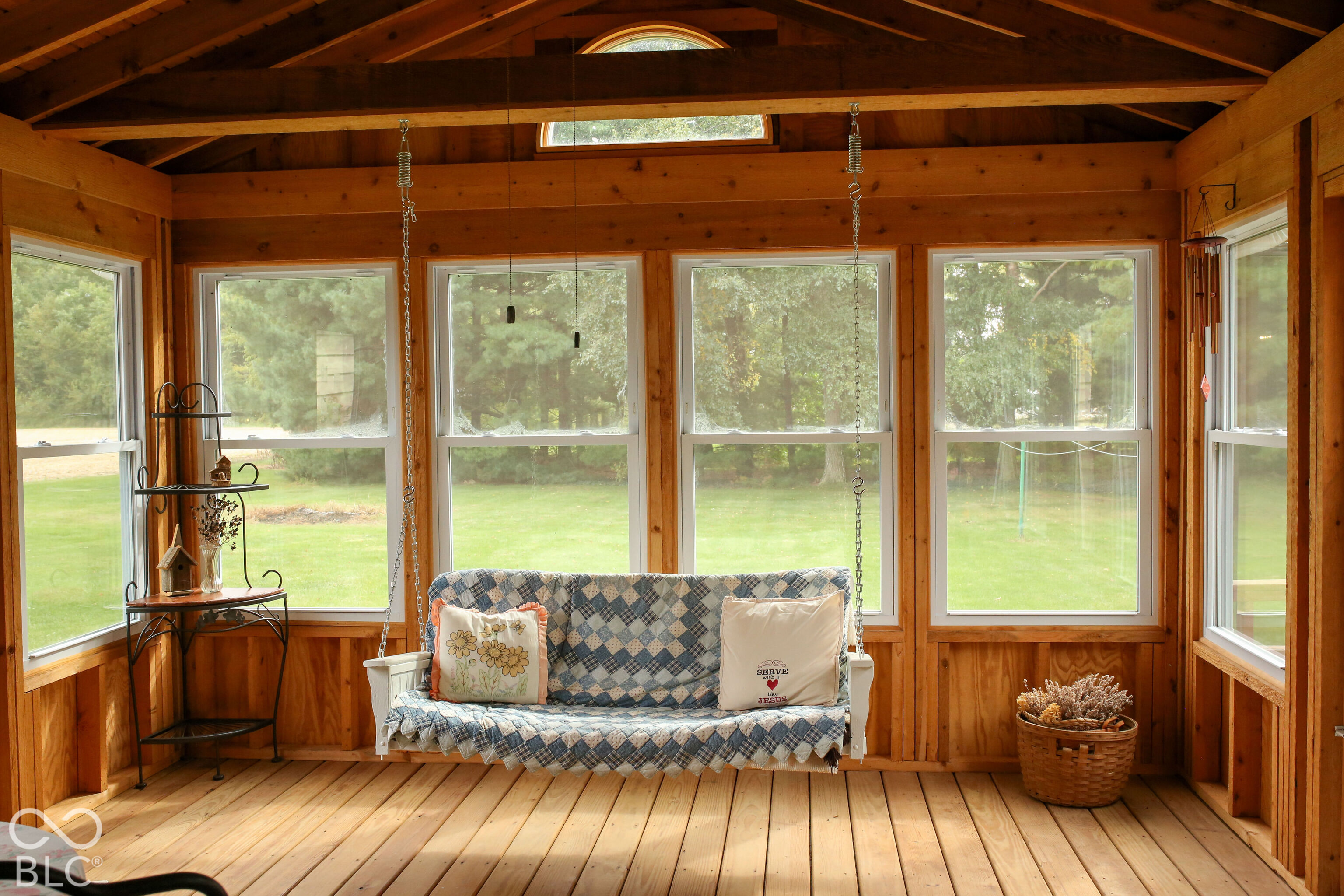 Wood-paneled sunroom with a quilt-covered hanging swing and large windows overlooking the lawn.