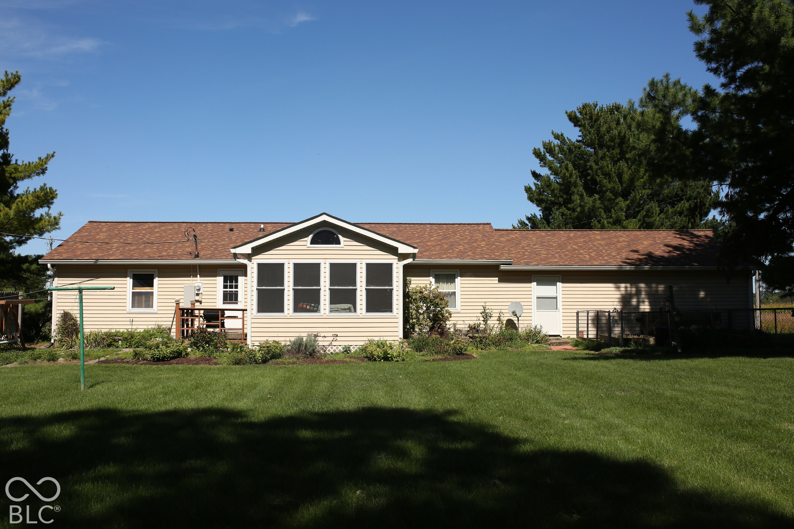 Rear of a beige ranch house with a central sunroom, broad lawn, and mature trees.