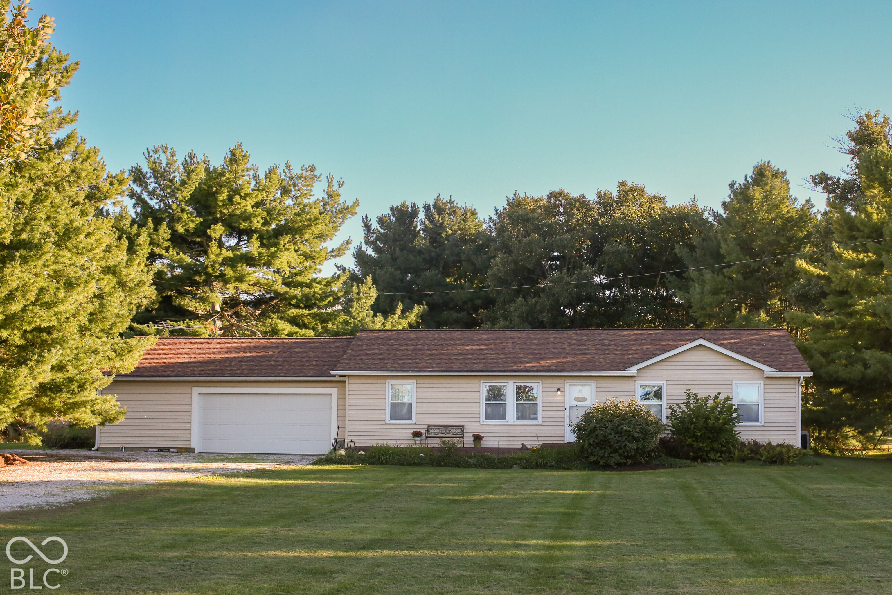 Single-story beige home with an attached garage, broad lawn, and mature trees.