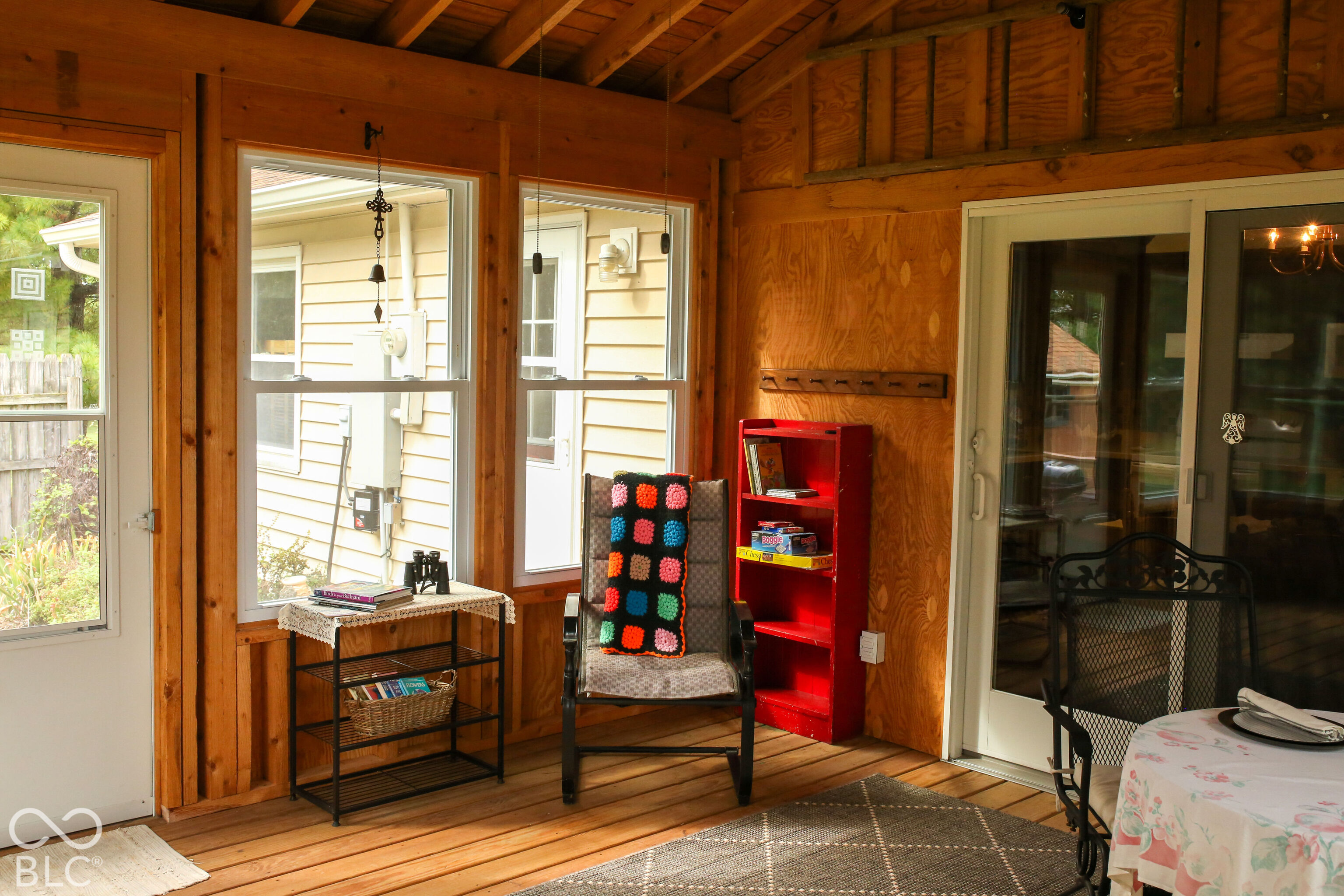 Enclosed porch with wood-paneled walls, large windows, a colorful throw, and a red bookcase.