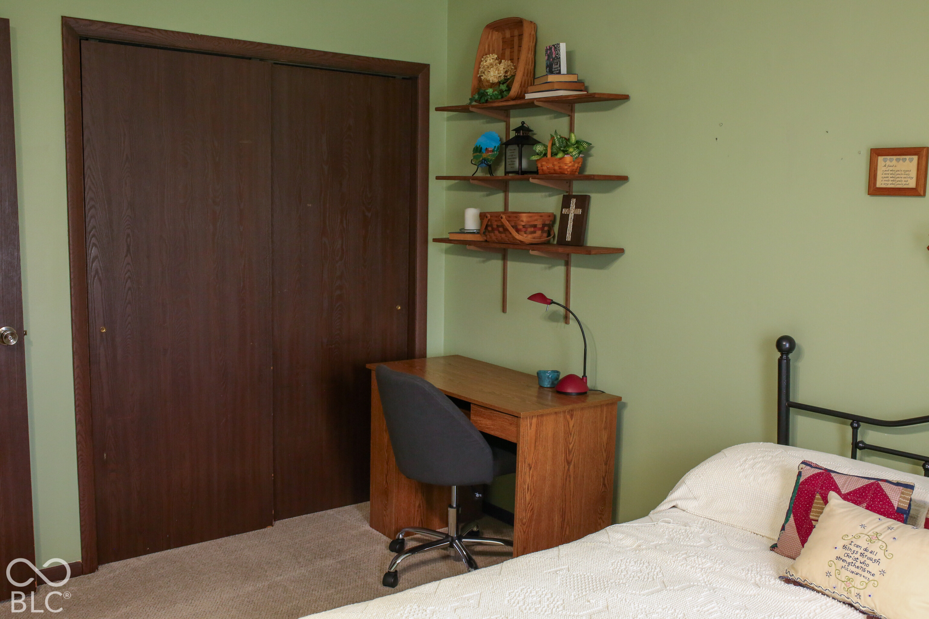 Bedroom with a white quilted bed, dark wood closet doors, and a desk beneath wall shelves.