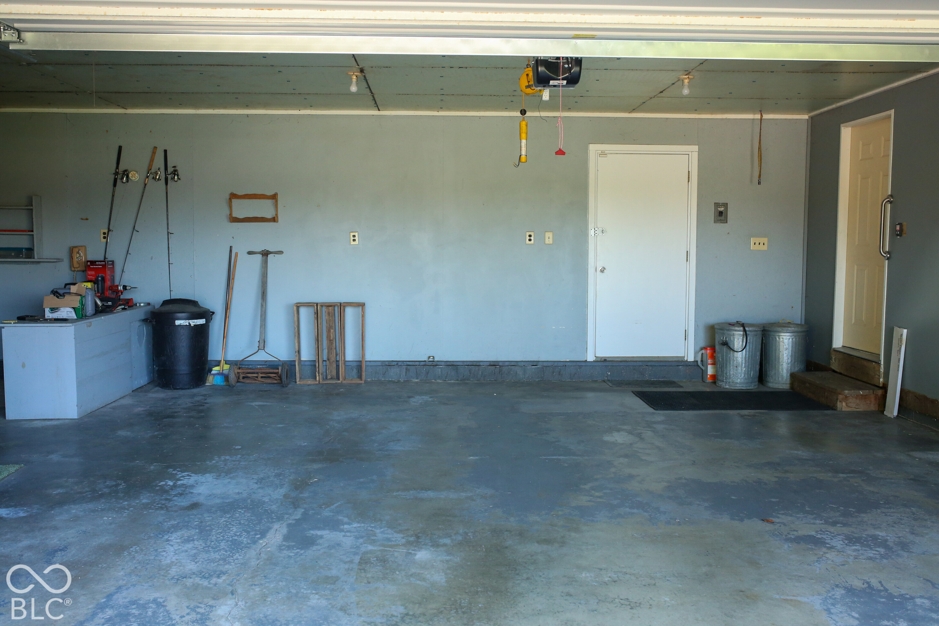 Garage with a broad concrete floor, overhead door opener, and tools and storage along the walls.