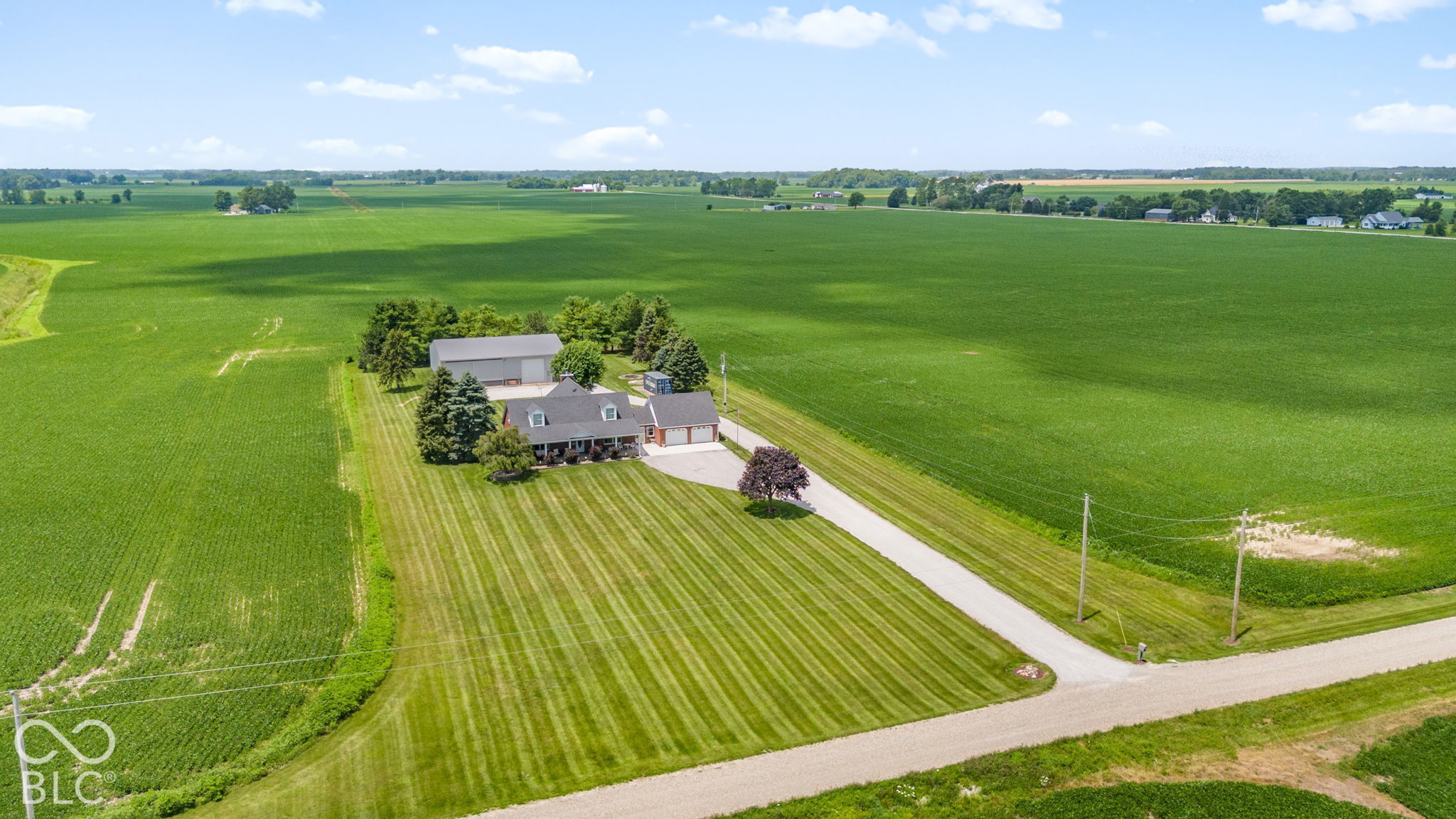 Rural home and outbuilding amid expansive green fields, with a long driveway and striped lawn.
