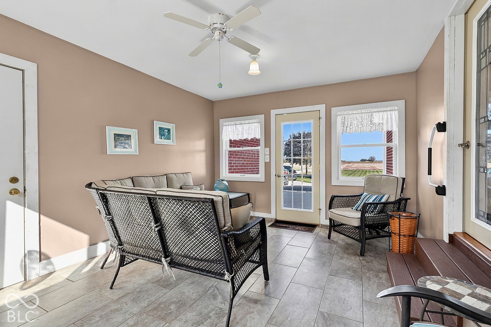 Furnished sunroom with wicker seating, gray tile floors, and windows overlooking a field.