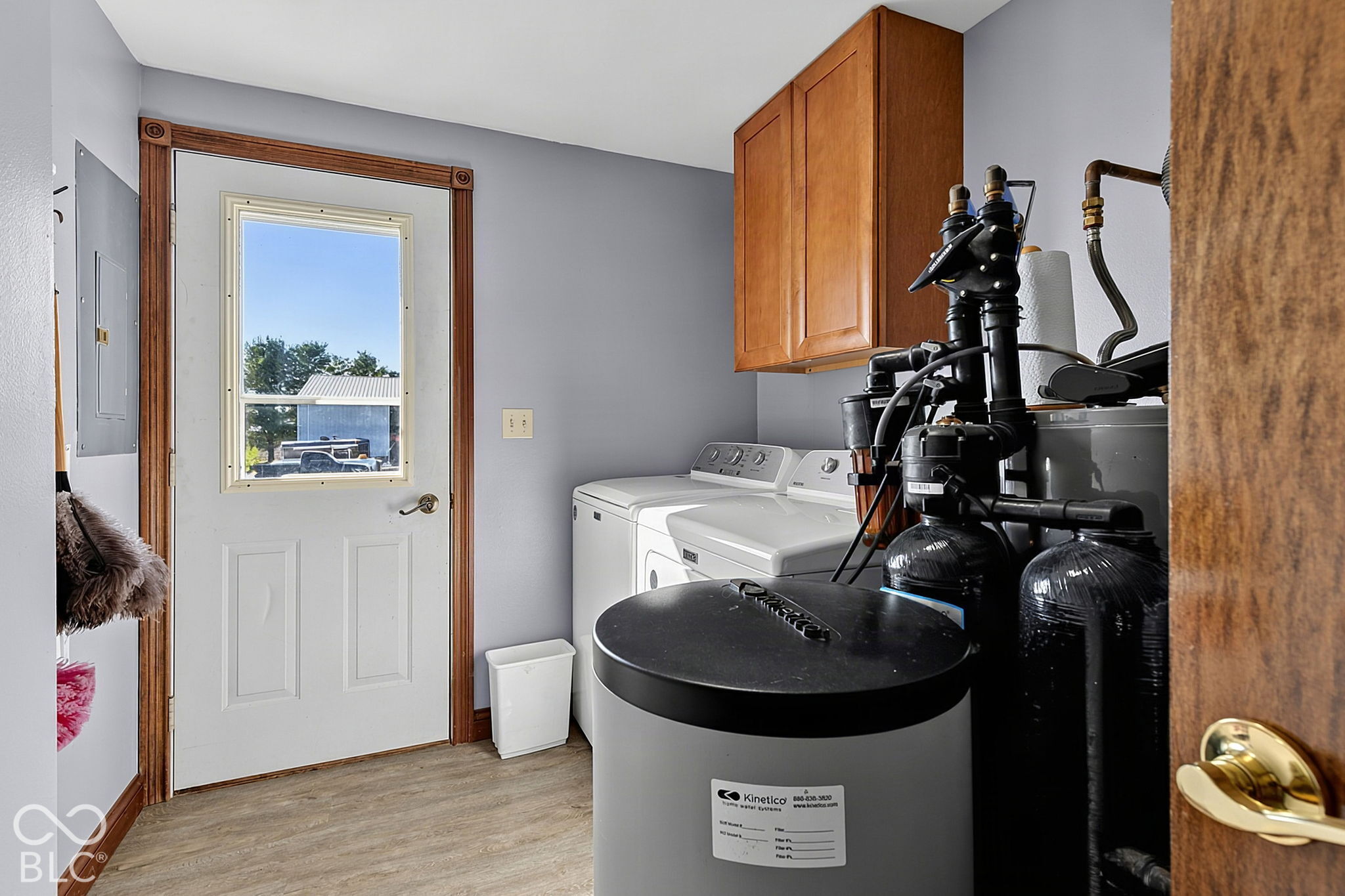 Laundry room with washer and dryer beside a large water softener system and wooden cabinets.