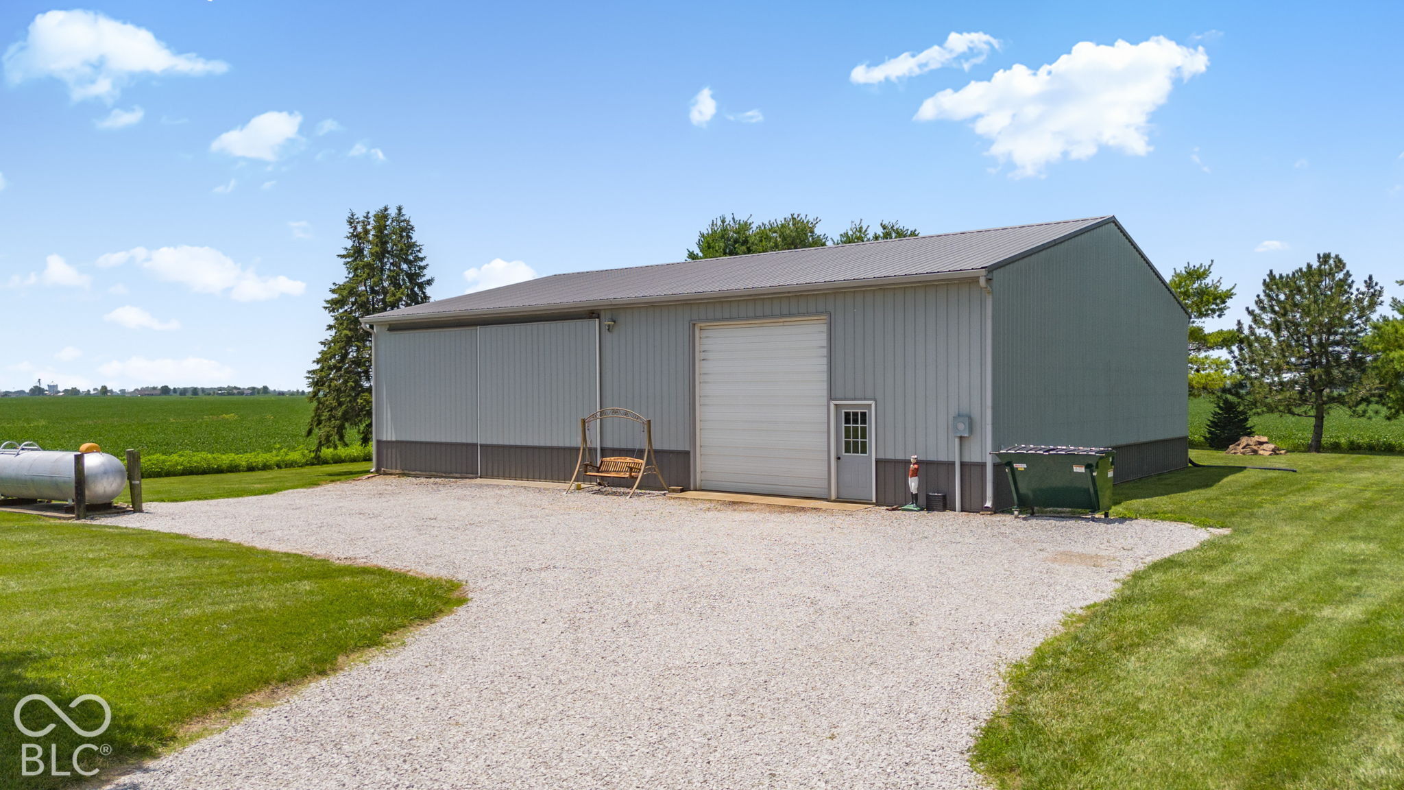 Large metal-sided barn with a wide overhead door beside a gravel drive and open fields.