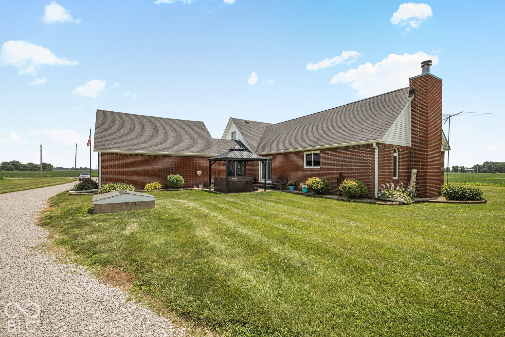 Red-brick house with a gazebo-covered hot tub on a broad lawn beside open fields.