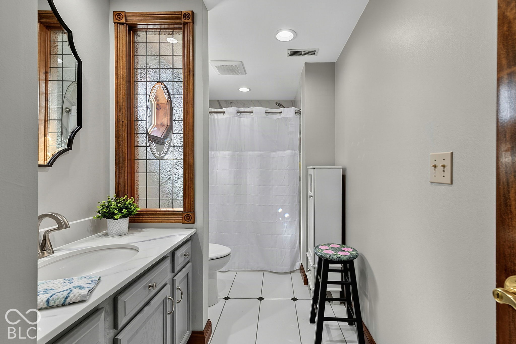 Bathroom with a gray vanity, decorative stained-glass window, curtained shower, and white tile floor.
