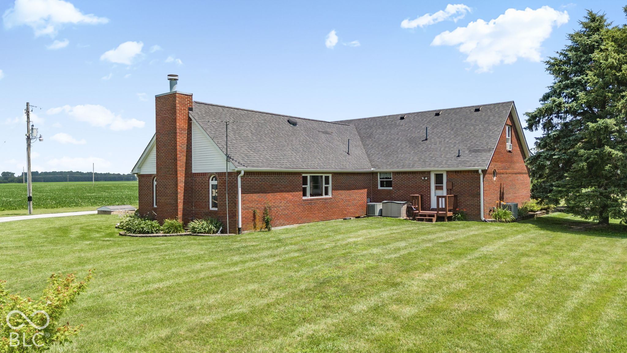 Red-brick house with a steep gray roof, small deck, and broad lawn beside farmland.