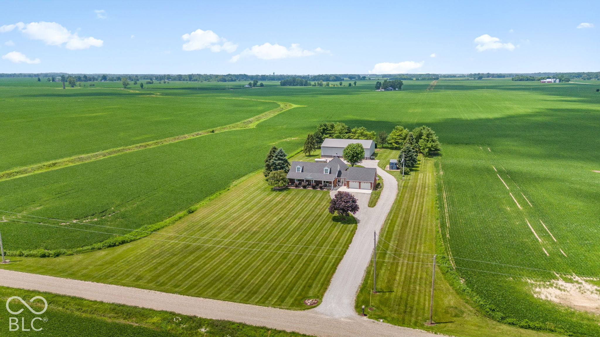 Aerial view of a rural home amid green fields, with a curving driveway and detached outbuilding.
