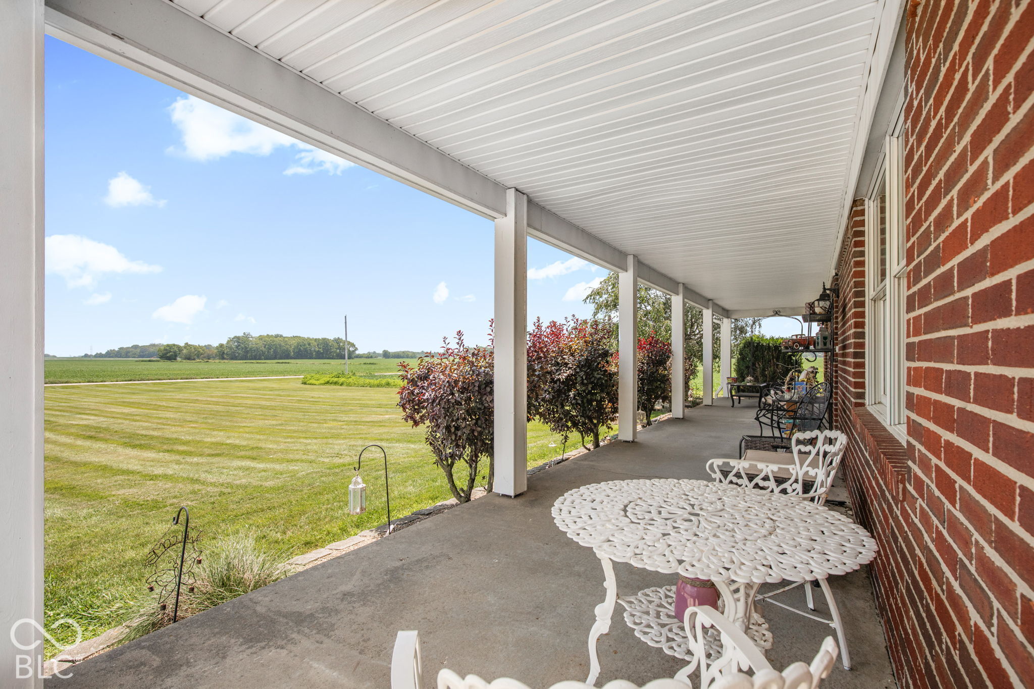 Covered brick-house porch with white patio furniture overlooking a broad lawn and open farmland.