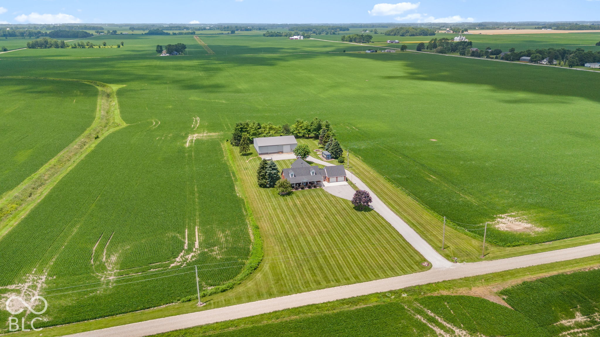Aerial view of a rural home and large outbuilding amid expansive green fields, with a long driveway.