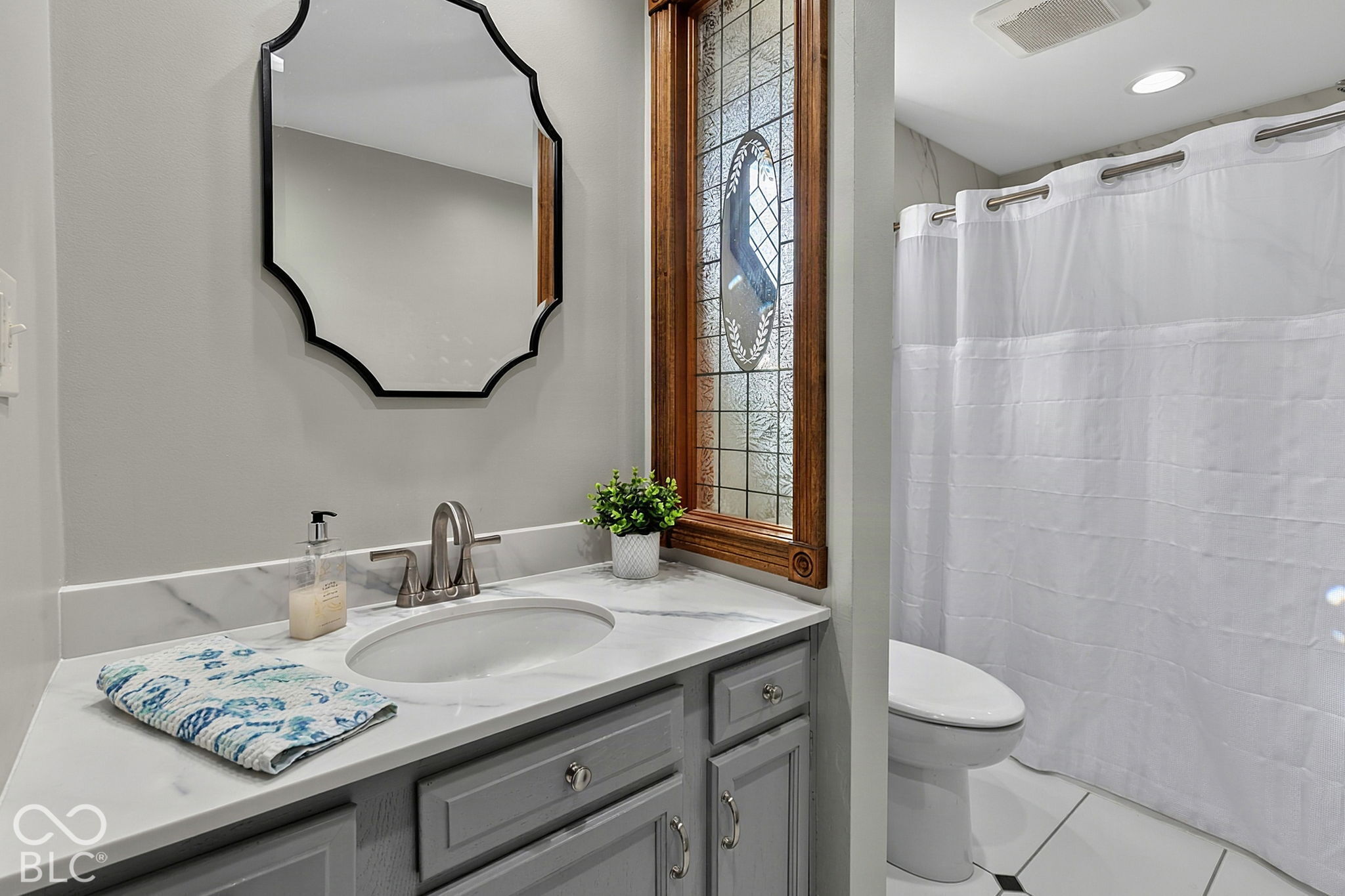 Bathroom with gray vanity, marble-look counter, and stained-glass window beside a curtained shower.