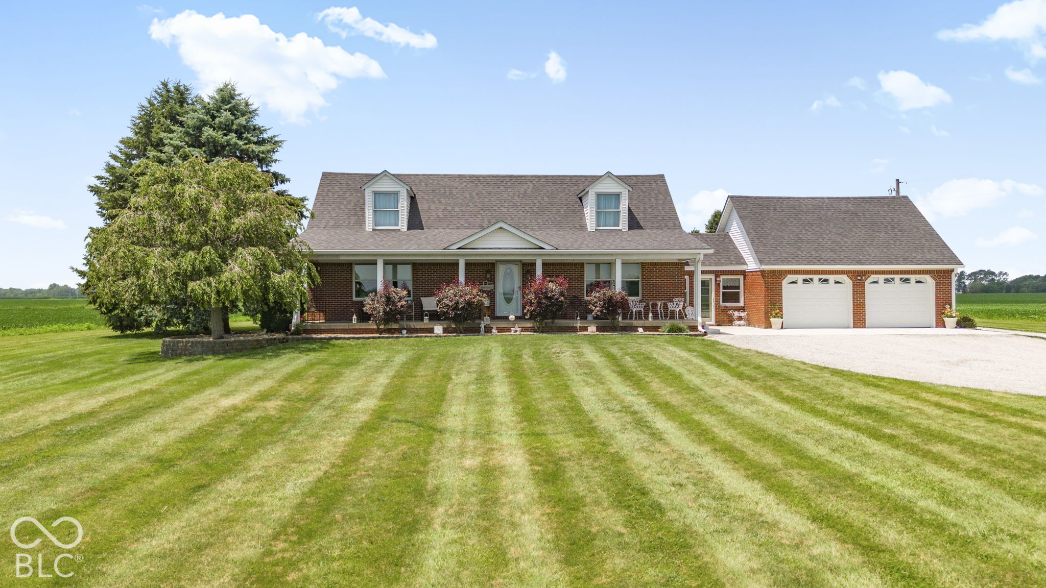 Brick ranch home with a covered porch, attached two-car garage, and broad lawn beside open fields.