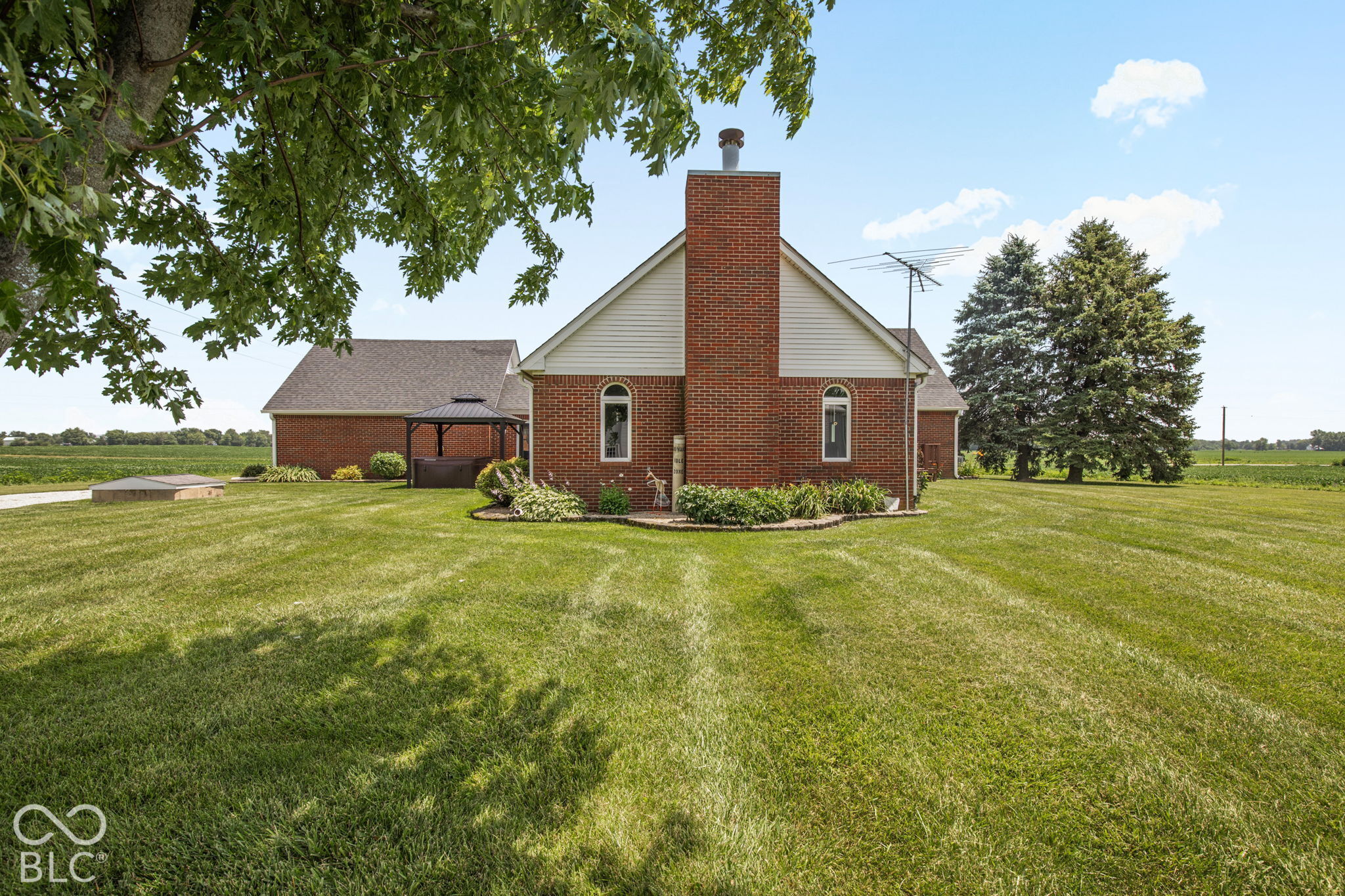 Red-brick house with a tall chimney, gazebo, and wide lawn beside open fields.
