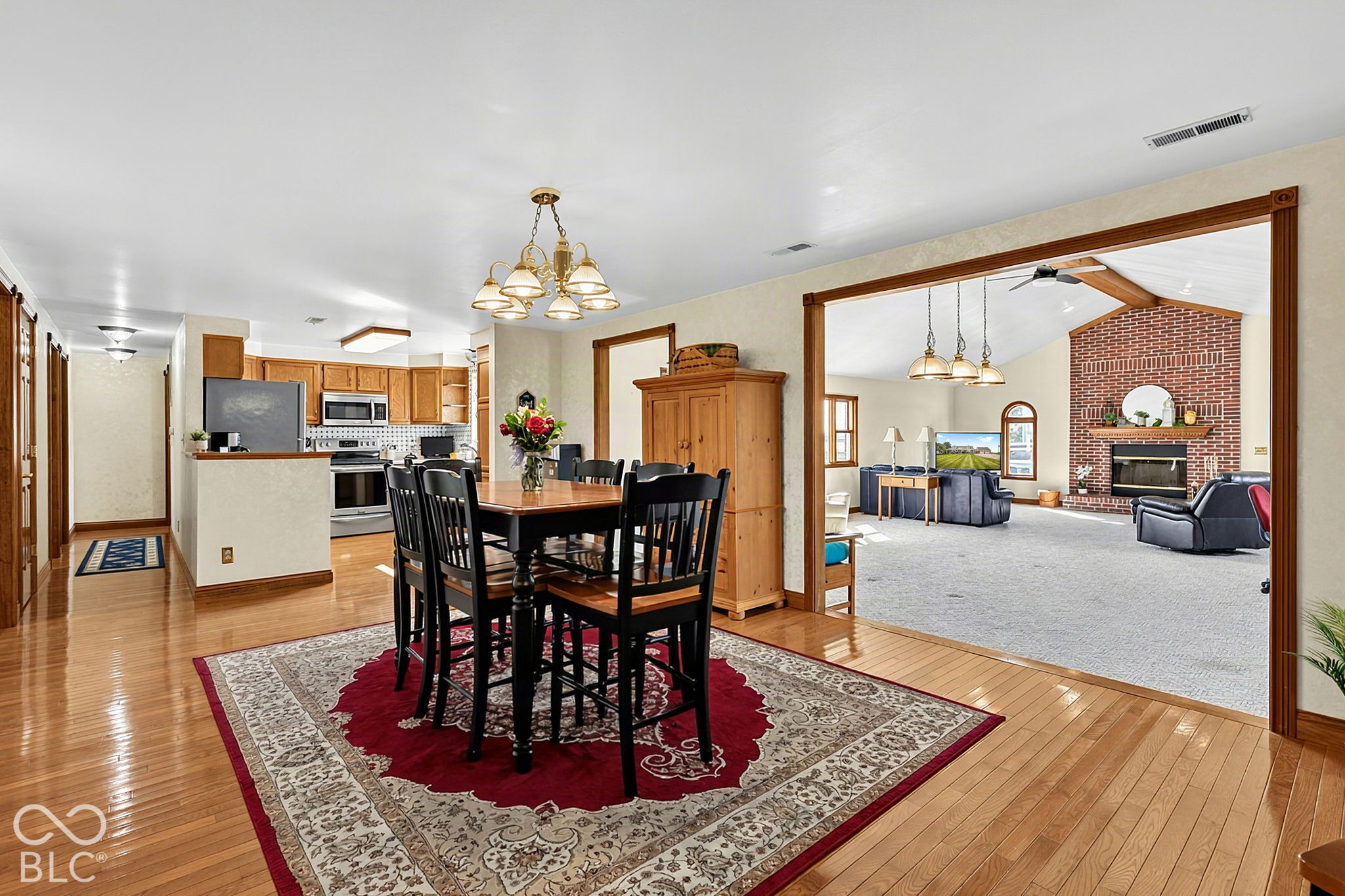 Dining room with a dark wood table and patterned rug, open to the kitchen and a living room with a brick fireplace.