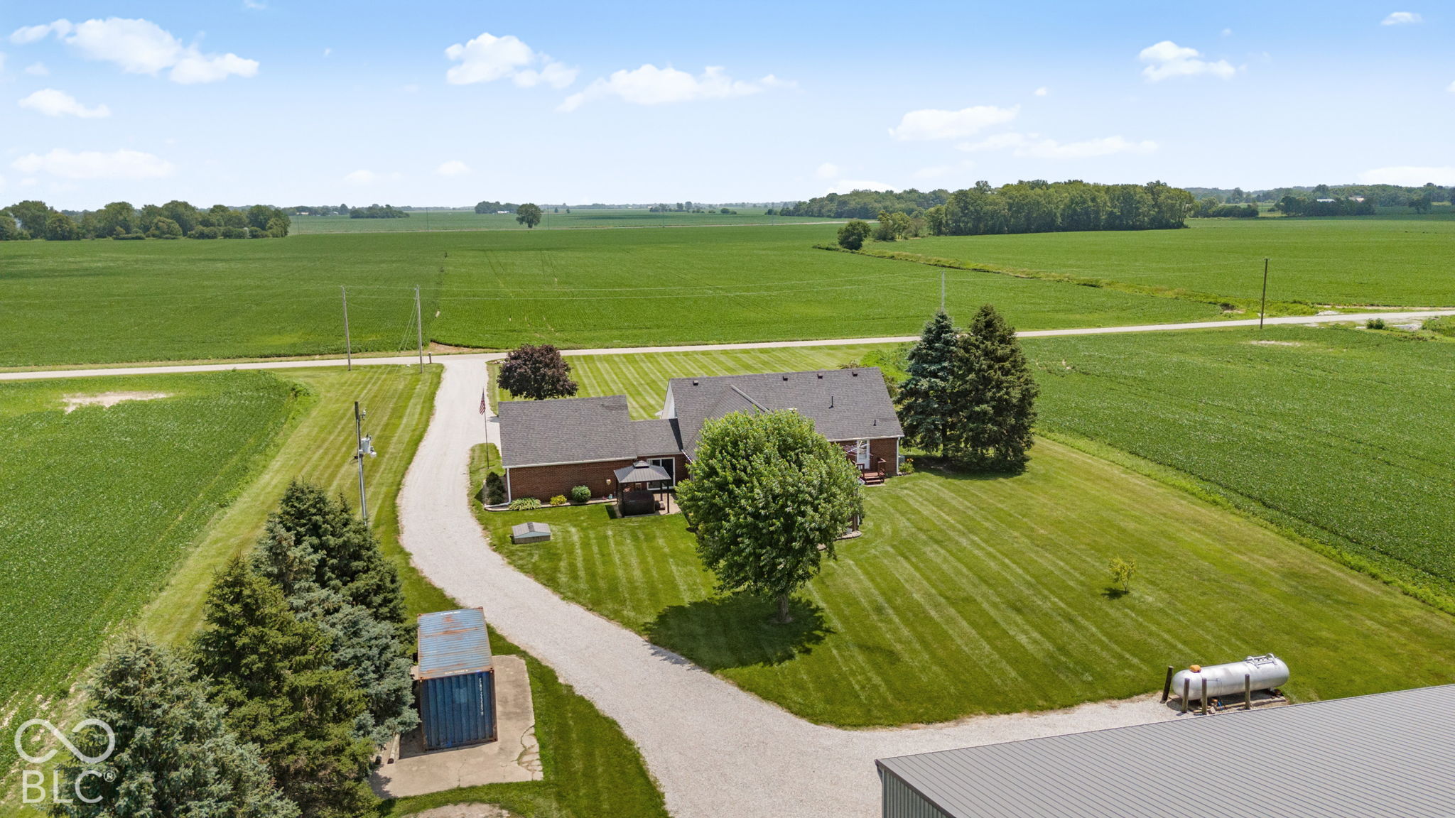 Aerial view of a rural home, gravel driveway, mowed lawn, and surrounding crop fields.
