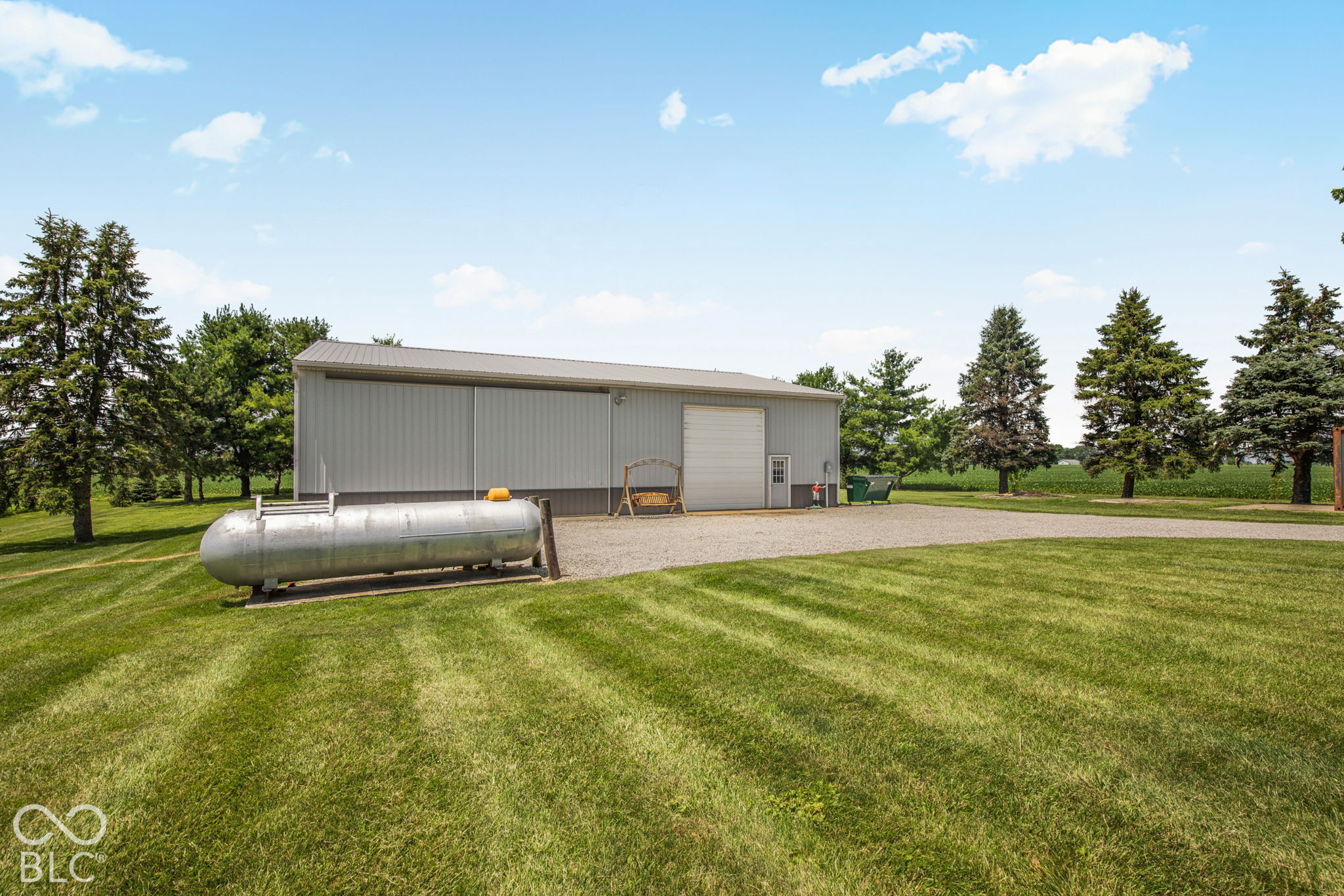 Gray metal pole barn with a roll-up door, gravel drive, and propane tank on a spacious lawn.