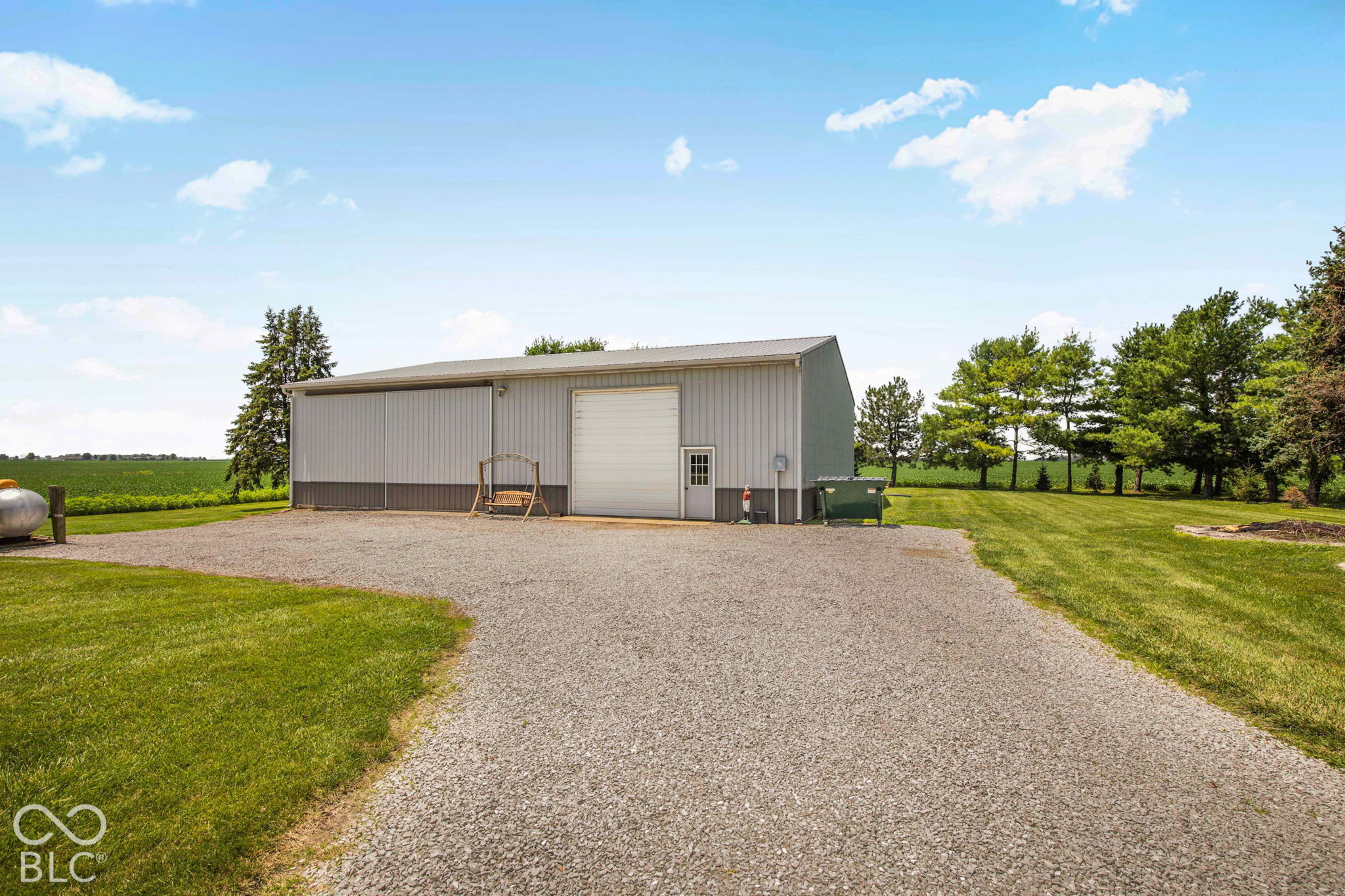 Gray metal outbuilding with a roll-up door beside a wide gravel driveway and open farmland.