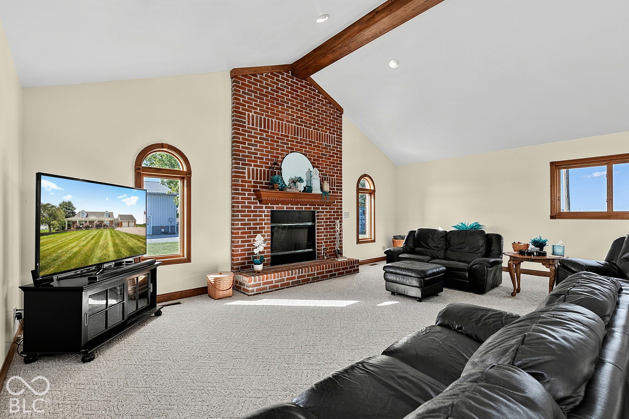 Living room with a tall brick fireplace, exposed ceiling beam, arched windows, and black leather seating.