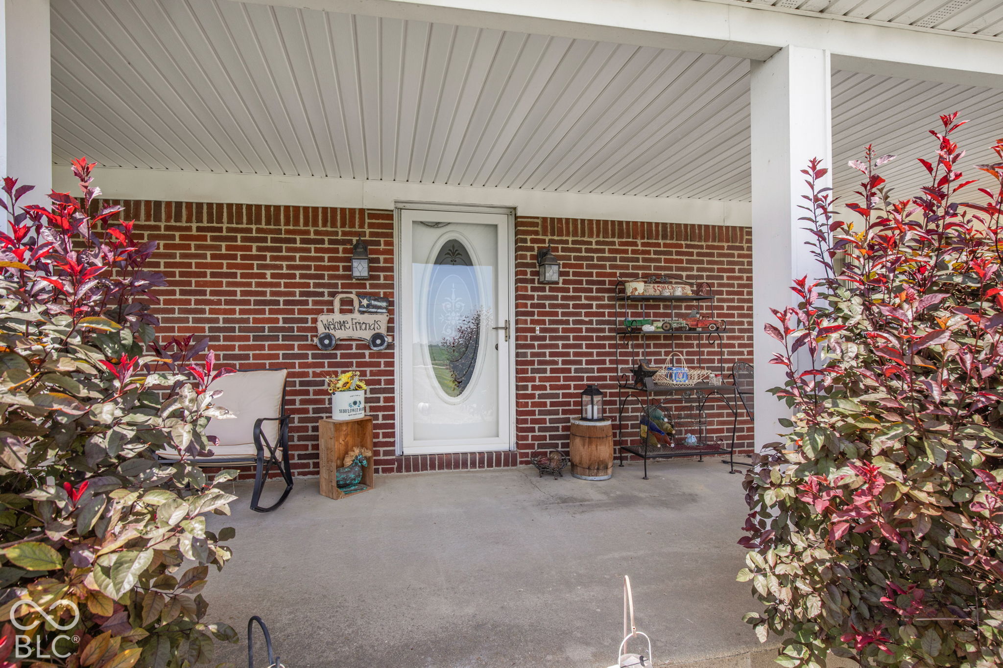 Covered brick front porch with a white oval-glass door, rocking chair, and red-leaf shrubs.