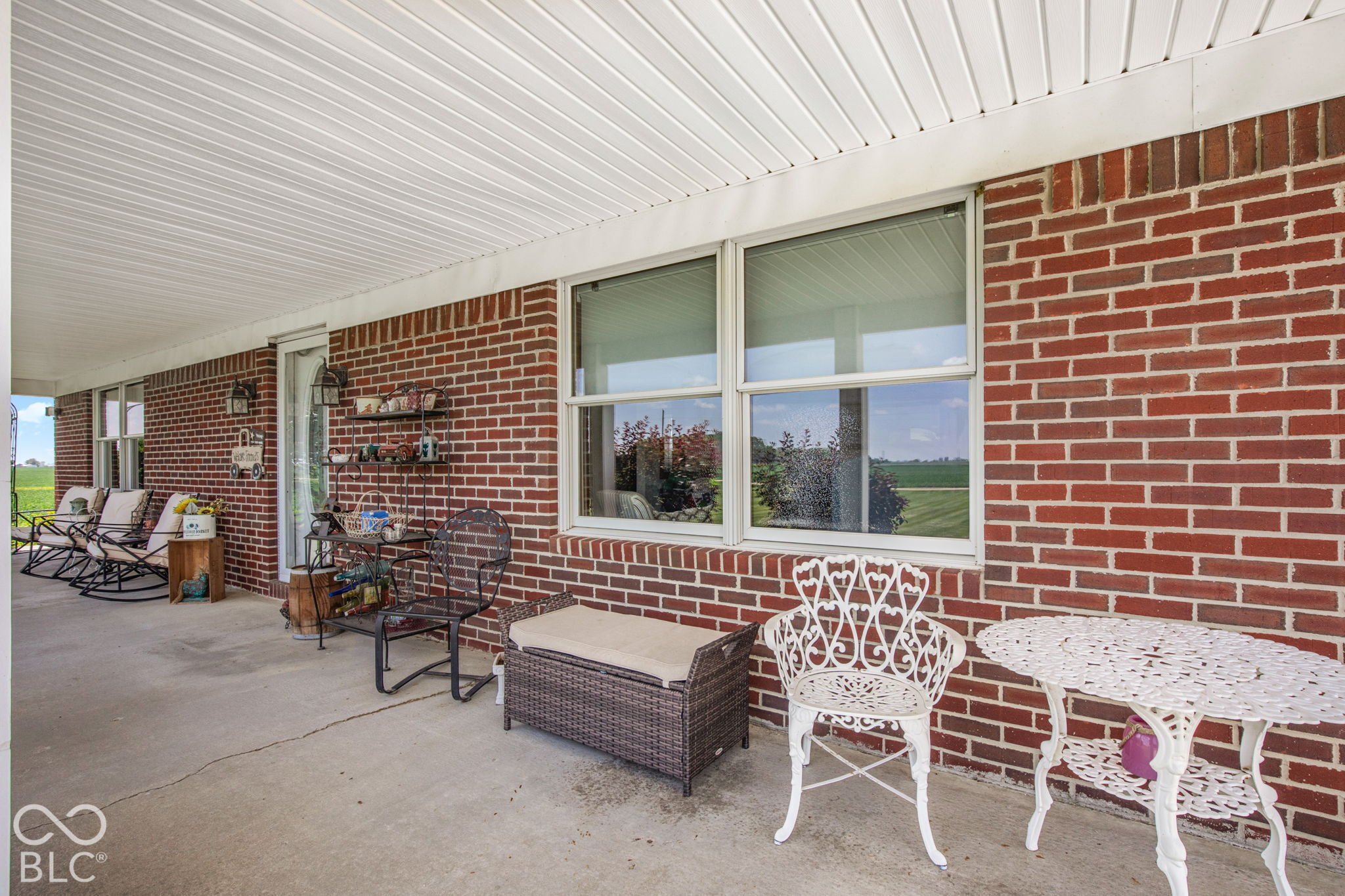 Covered brick patio with white metal furniture, a cushioned bench, and open-field views.