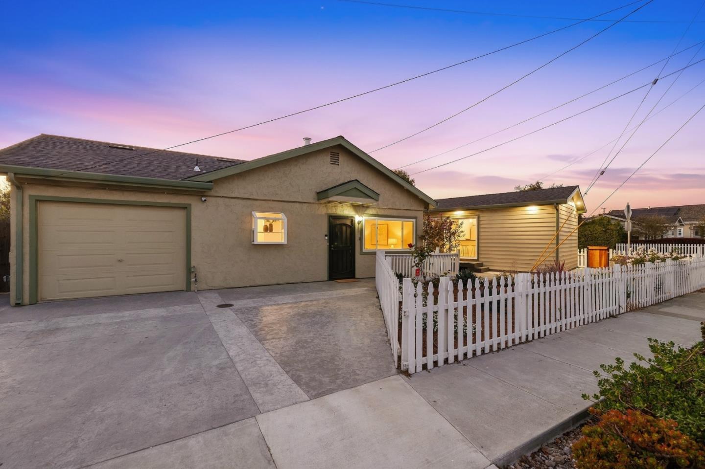 Single-story beige home with illuminated windows, an attached garage, and a white picket fence at dusk.