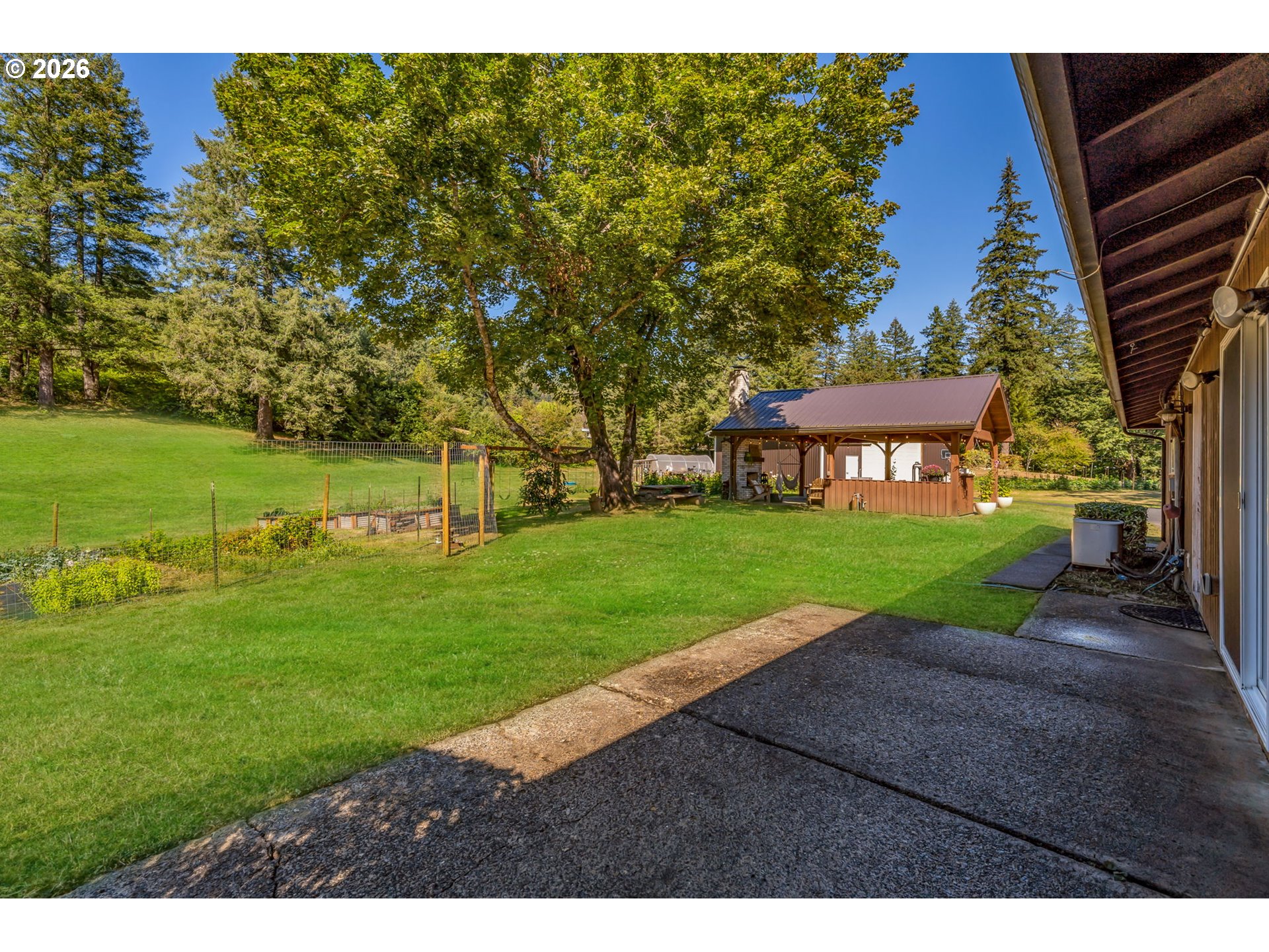 Backyard with a broad lawn, mature trees, fenced garden beds, and a covered pavilion beside the patio.