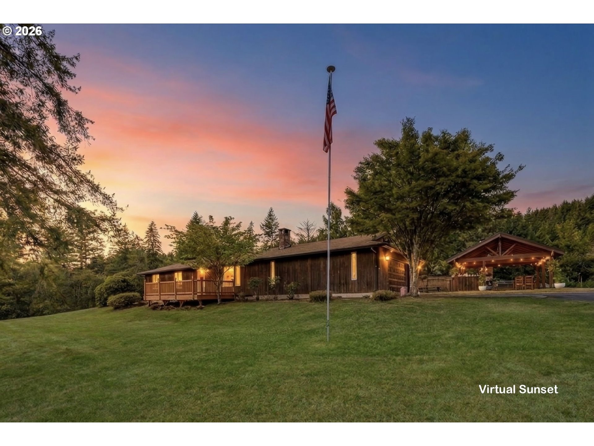 Wood-sided home and covered pavilion on a tree-lined lawn beneath a pink sunset sky.