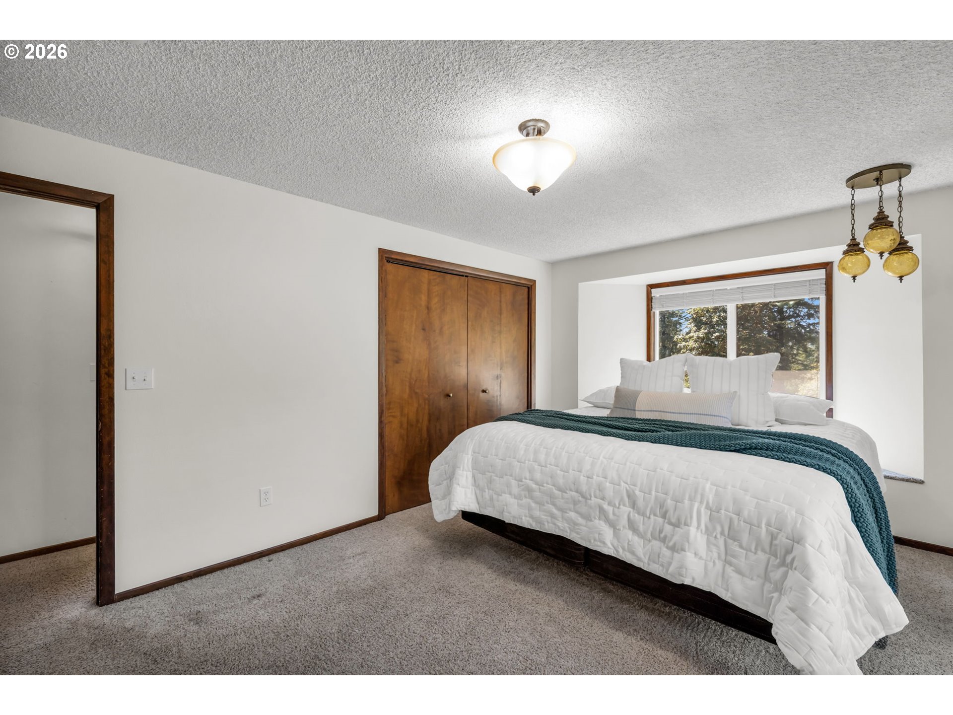 Bedroom with a white quilted bed, teal throw, wood closet doors, and a window overlooking trees.