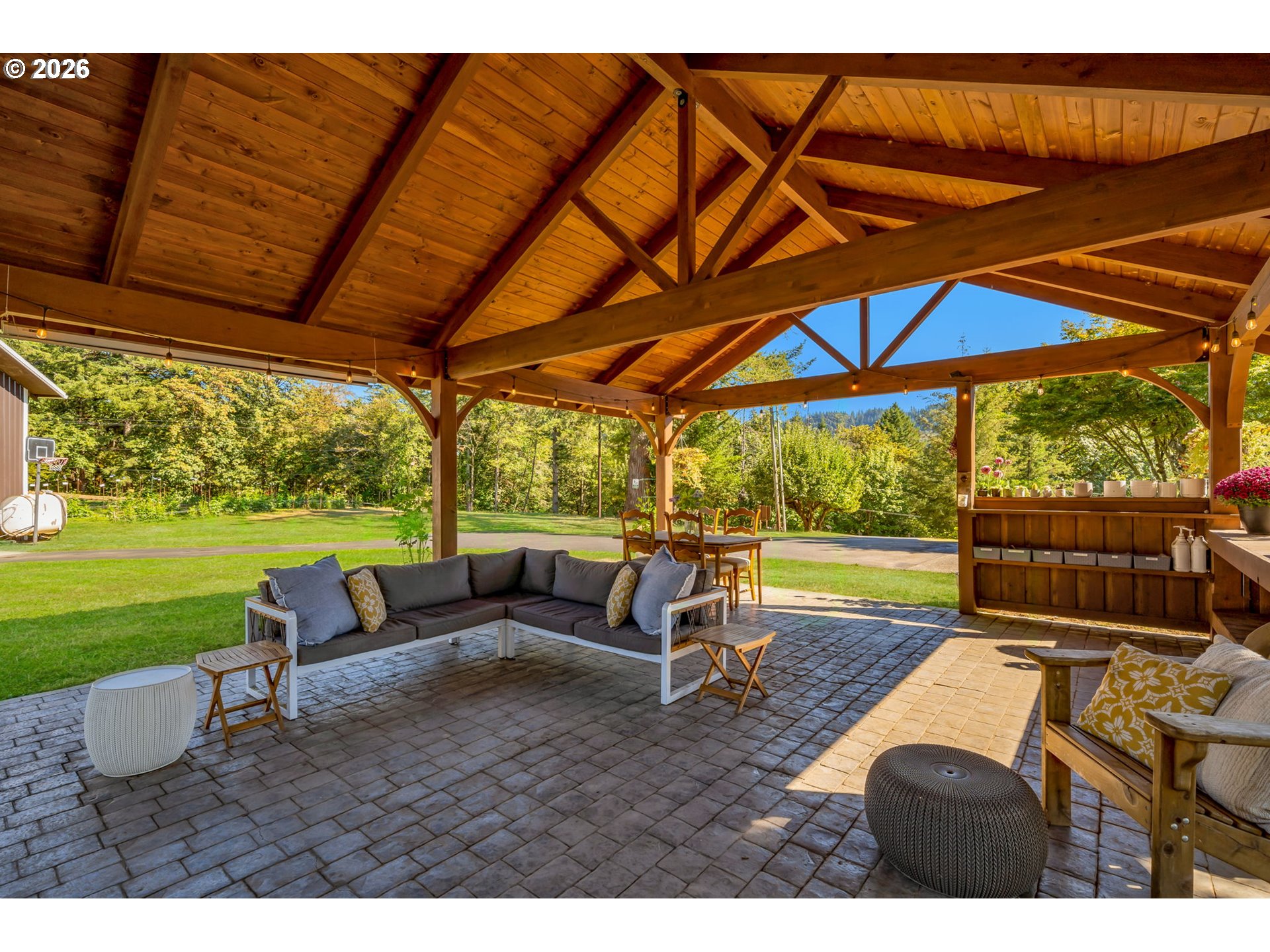 Covered timber patio with a sectional sofa, dining area, and views of a green lawn and trees.