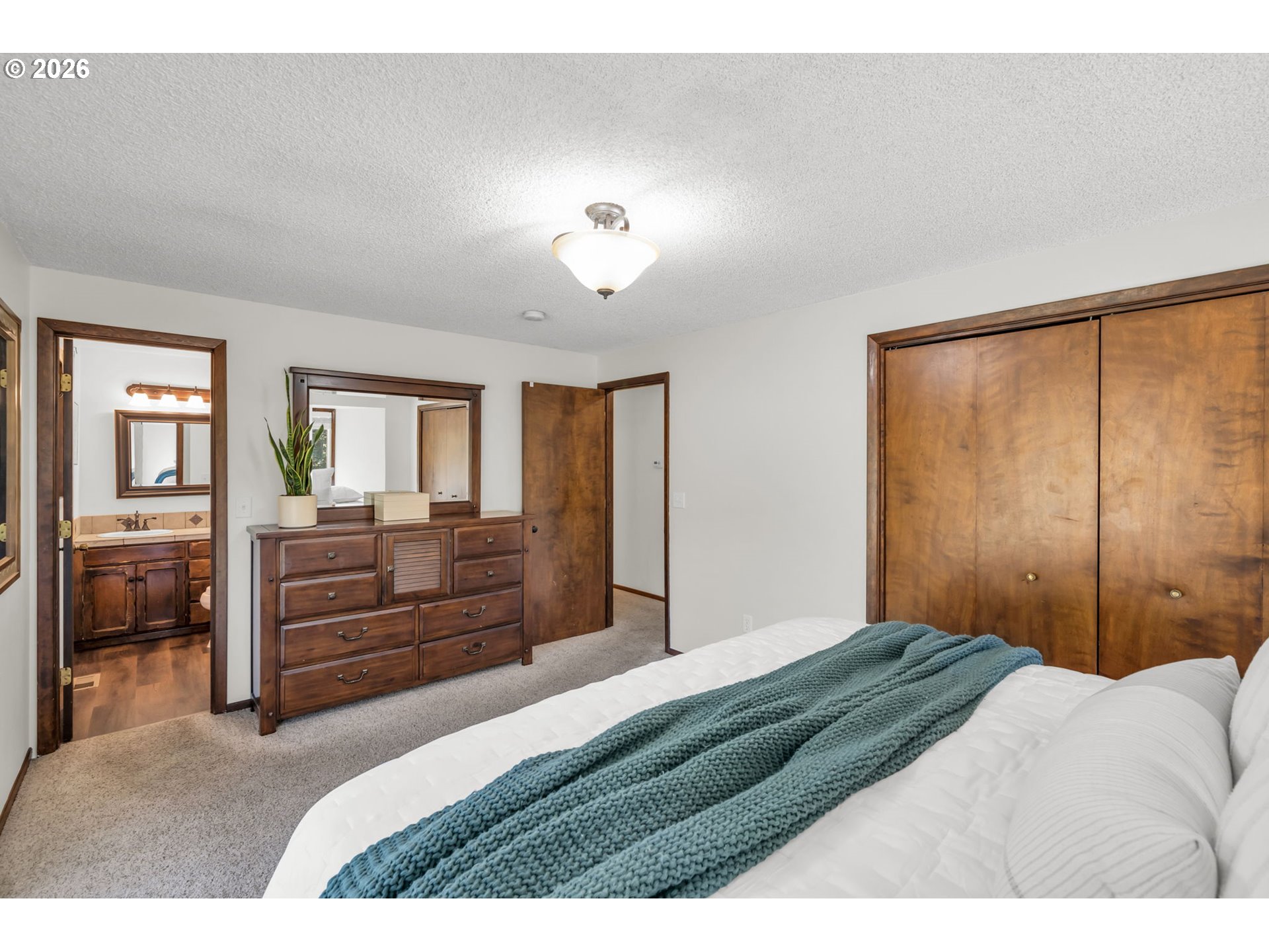 Bedroom with a teal throw, wood closet doors, and a dresser beside an open bathroom doorway.