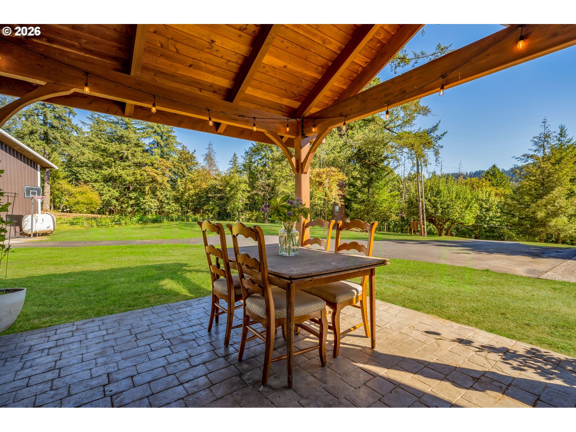 Covered timber patio with a dining table and chairs overlooking a green lawn and wooded yard.