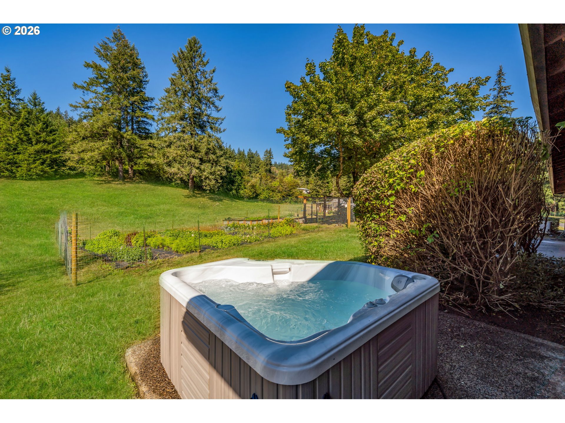Outdoor hot tub beside a grassy yard and fenced garden, backed by mature trees.